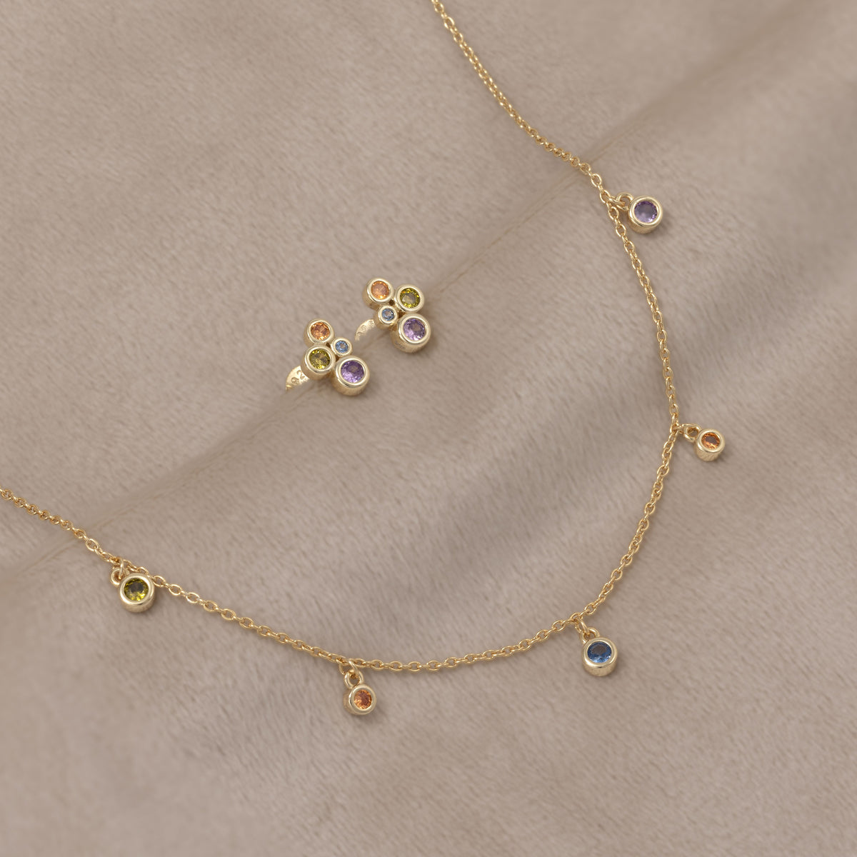 Pair of gold vermeil stud earrings featuring clusters of colourful purple and green bubble-shaped gemstones, presented alongside a delicate matching chain necklace with vibrant gem accents. Placed on a cream velvet cloth.