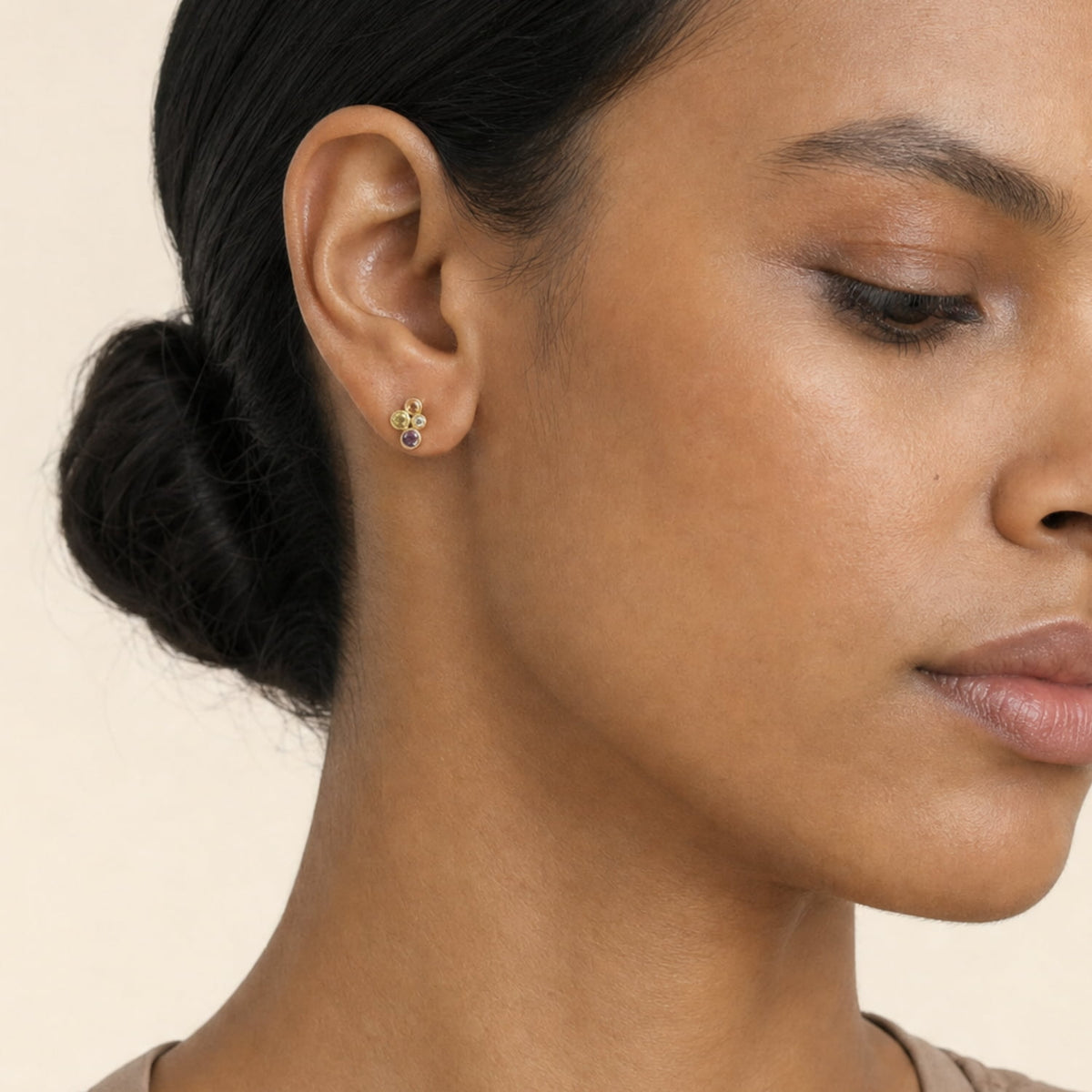 A close-up of a woman's ear wearing a small gold vermeil stud earring with acolourful cubic zirconia cluster, against a plain beige background.