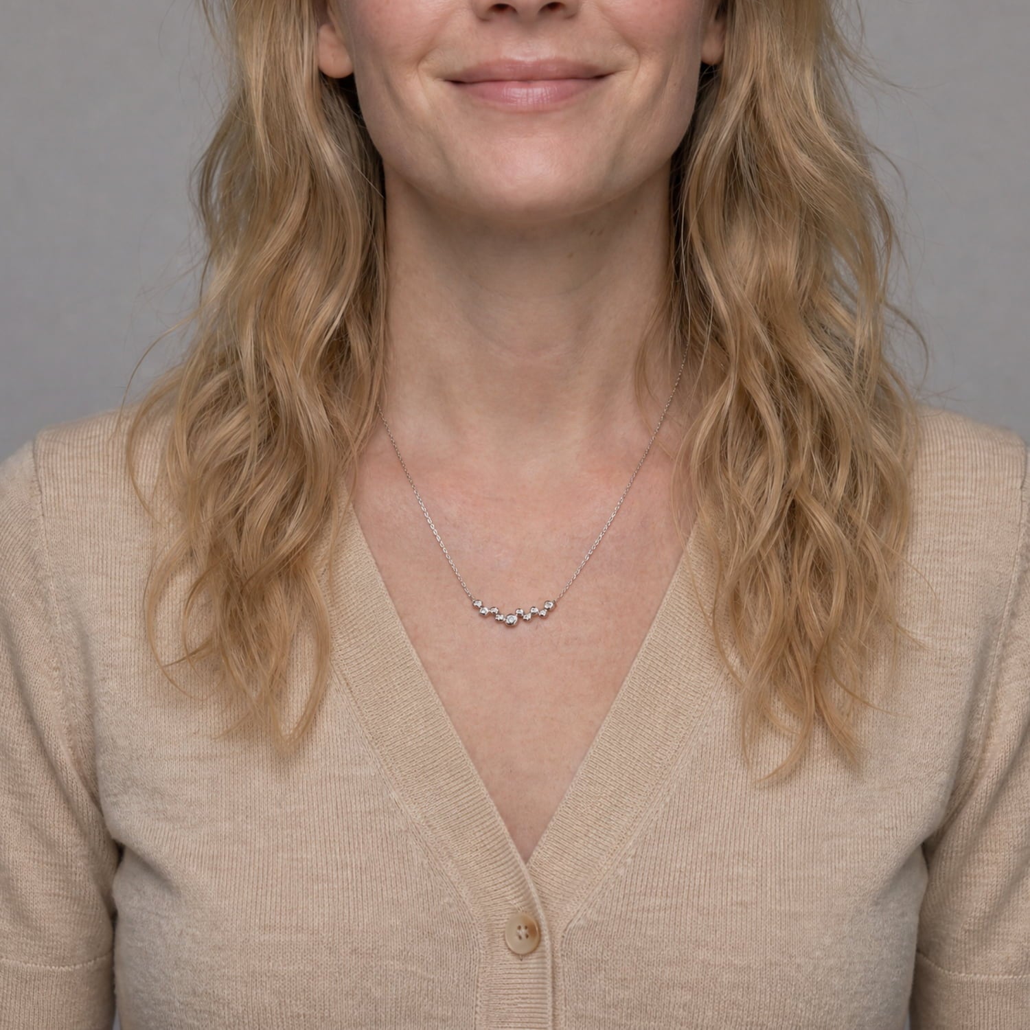A close-up of a woman wearing a delicate silver necklace with a small curved cluster of clear stones, styled with a beige cardigan against a neutral gray background.