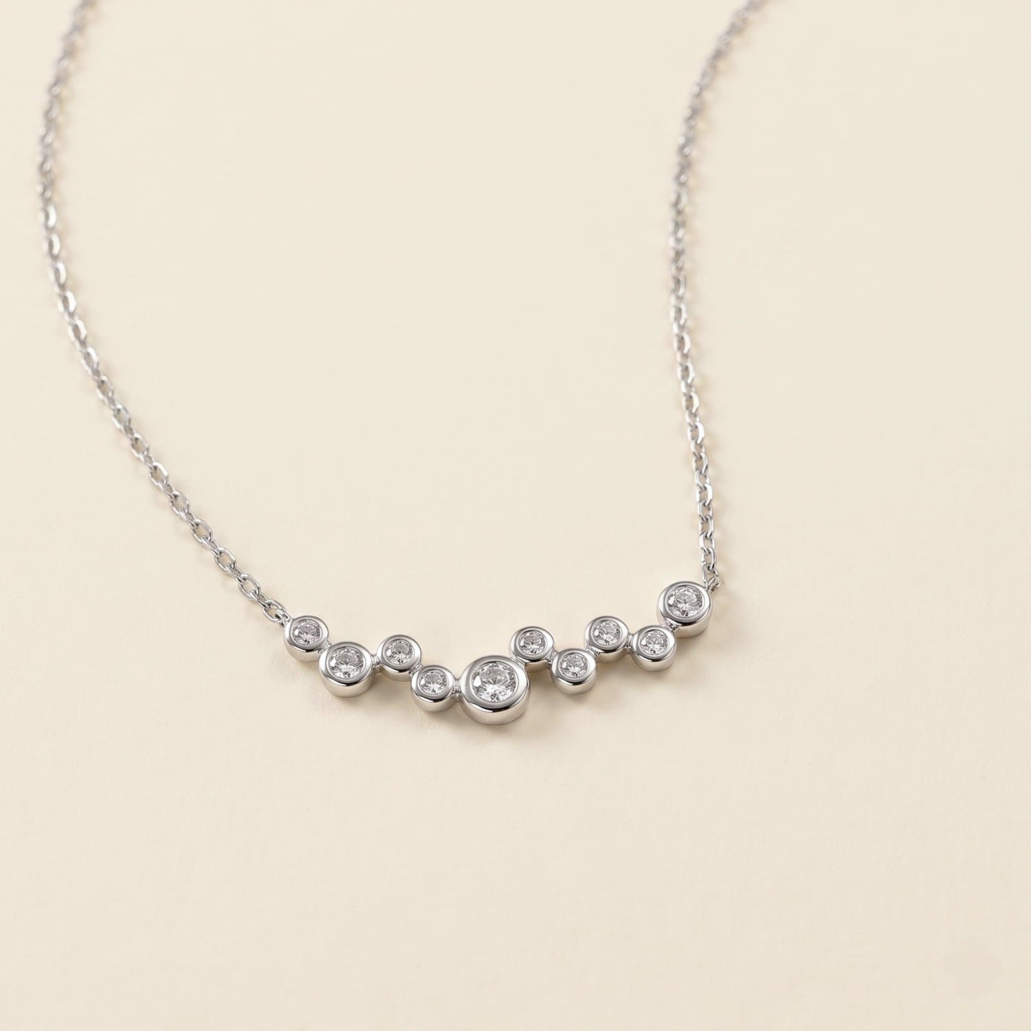 A delicate silver necklace with a curved bar of small clear cubic zirconia, displayed on a warm cream background.