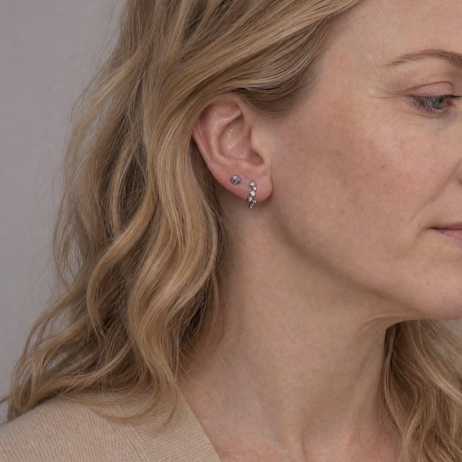 A close-up of a woman's ear wearing a pair of small silver half-hoop stud earrings with clear stones, shown against a soft neutral background.