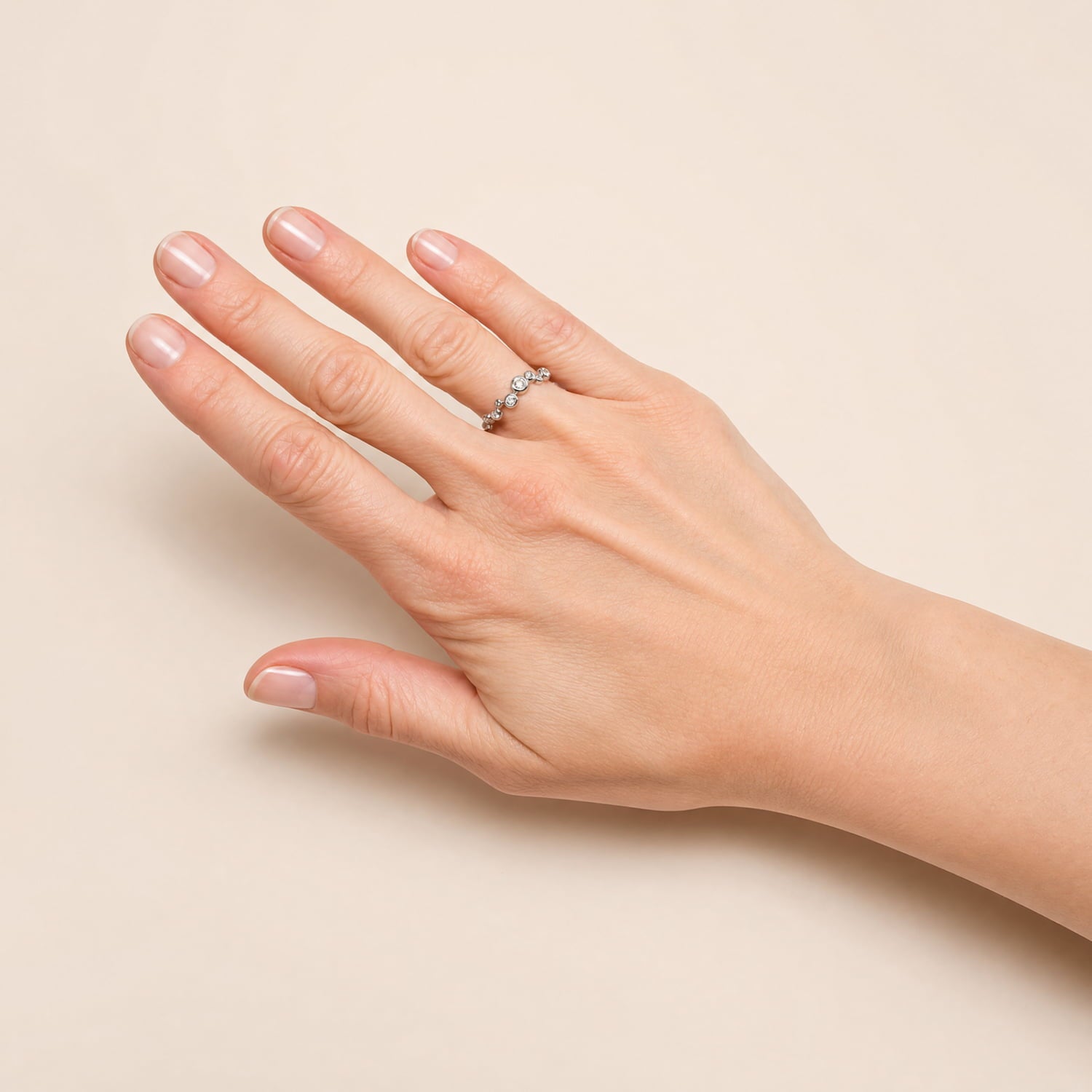 A close-up of a person's hand wearing a delicate silver ring with small clear stones, set against a solid light beige background.