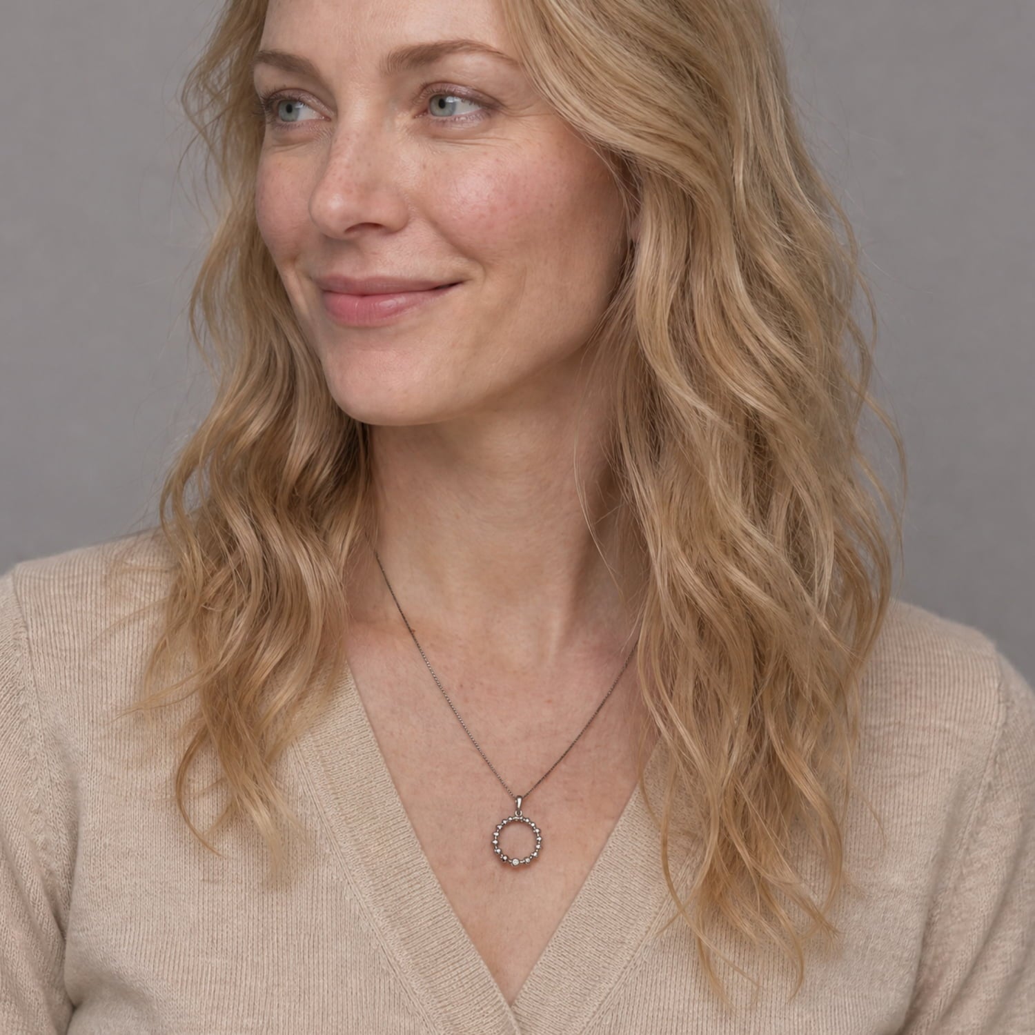 A woman with blonde wavy hair wearing a silver circular pendant necklace with a small clear stone, paired with a beige knit V-neck sweater.