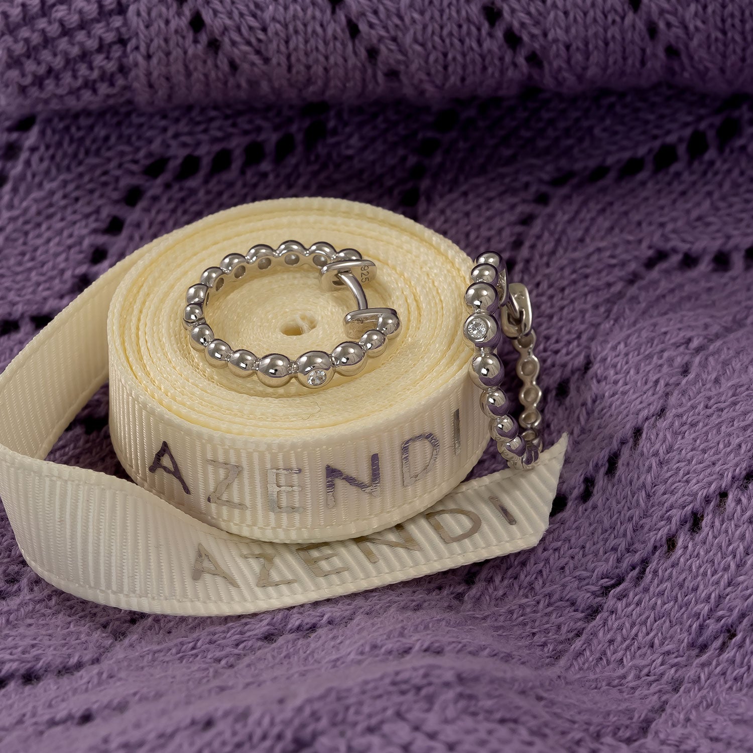 Pair of sterling silver hoop earrings featuring graduated bead detailing with cubic zirconia stones, displayed on a folded cream ribbon branded with Azendi, set against a textured purple fabric.