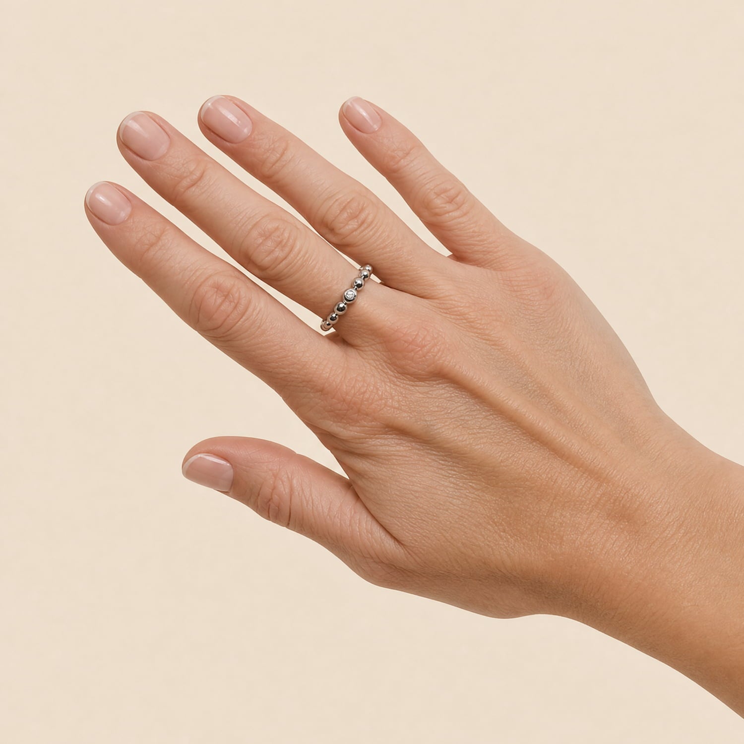 A close-up of a hand wearing a delicate silver ring with a small clear stone, set against a solid light beige background.
