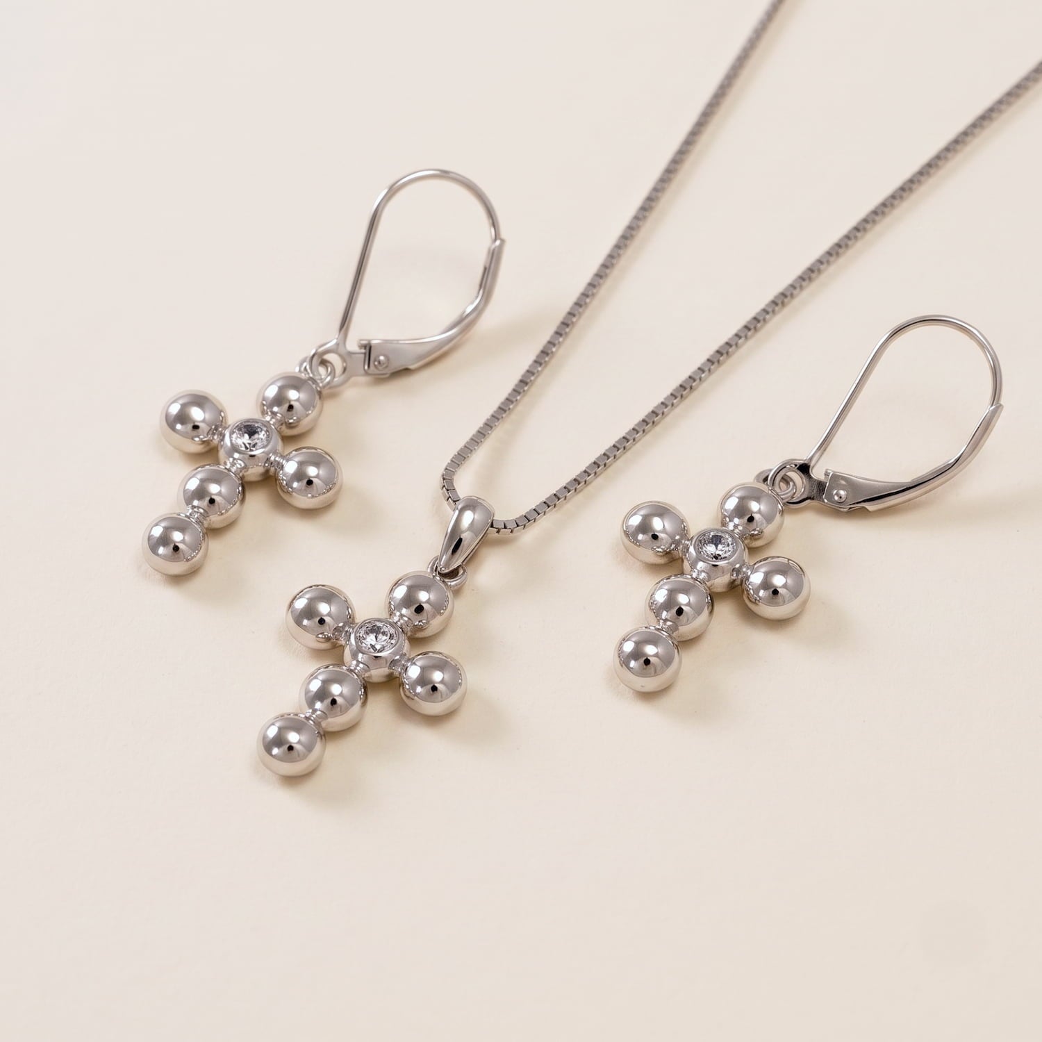 Sterling silver drop earrings and matching pendant featuring sculptural beaded cross designs accented with sparkling cubic zirconia stones, displayed on a cream surface.