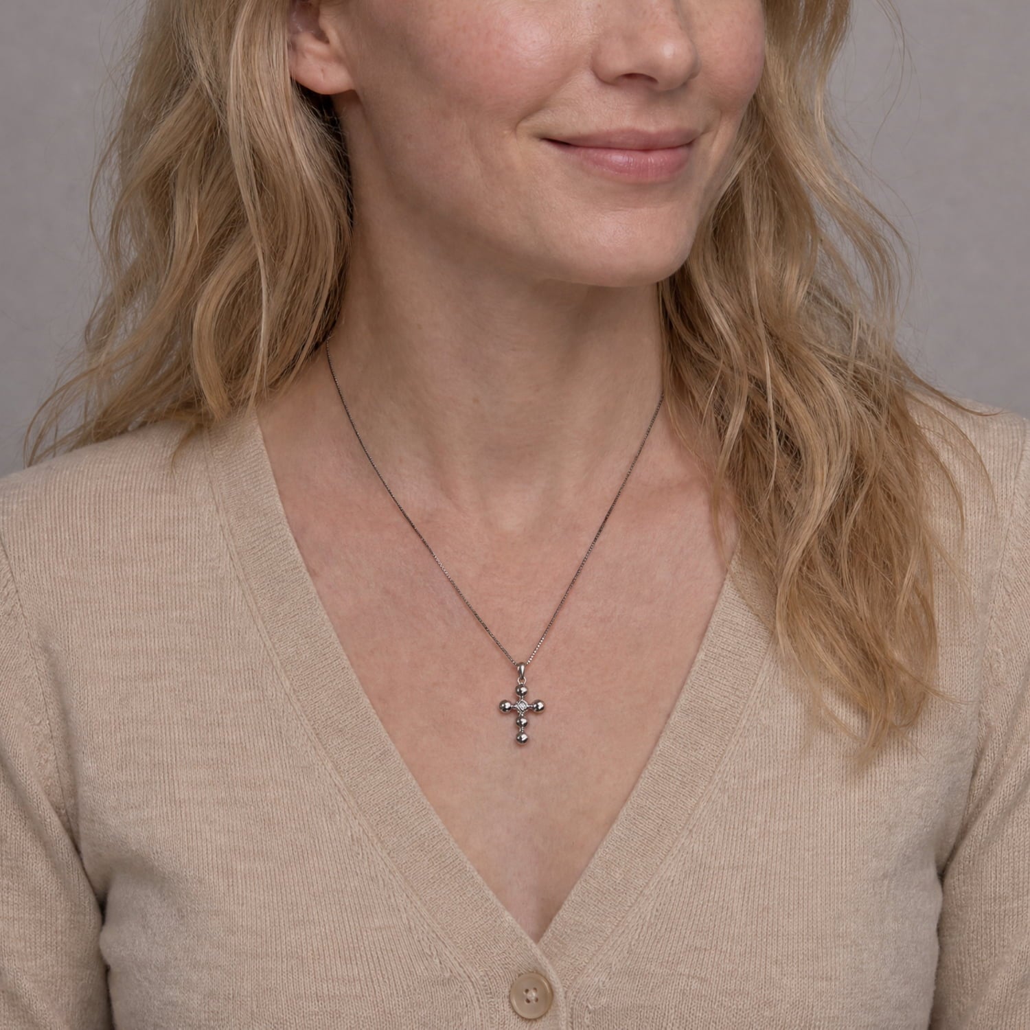 A close-up of a woman wearing a delicate silver cross pendant necklace with small clear stones, styled with a beige knit V-neck cardigan.