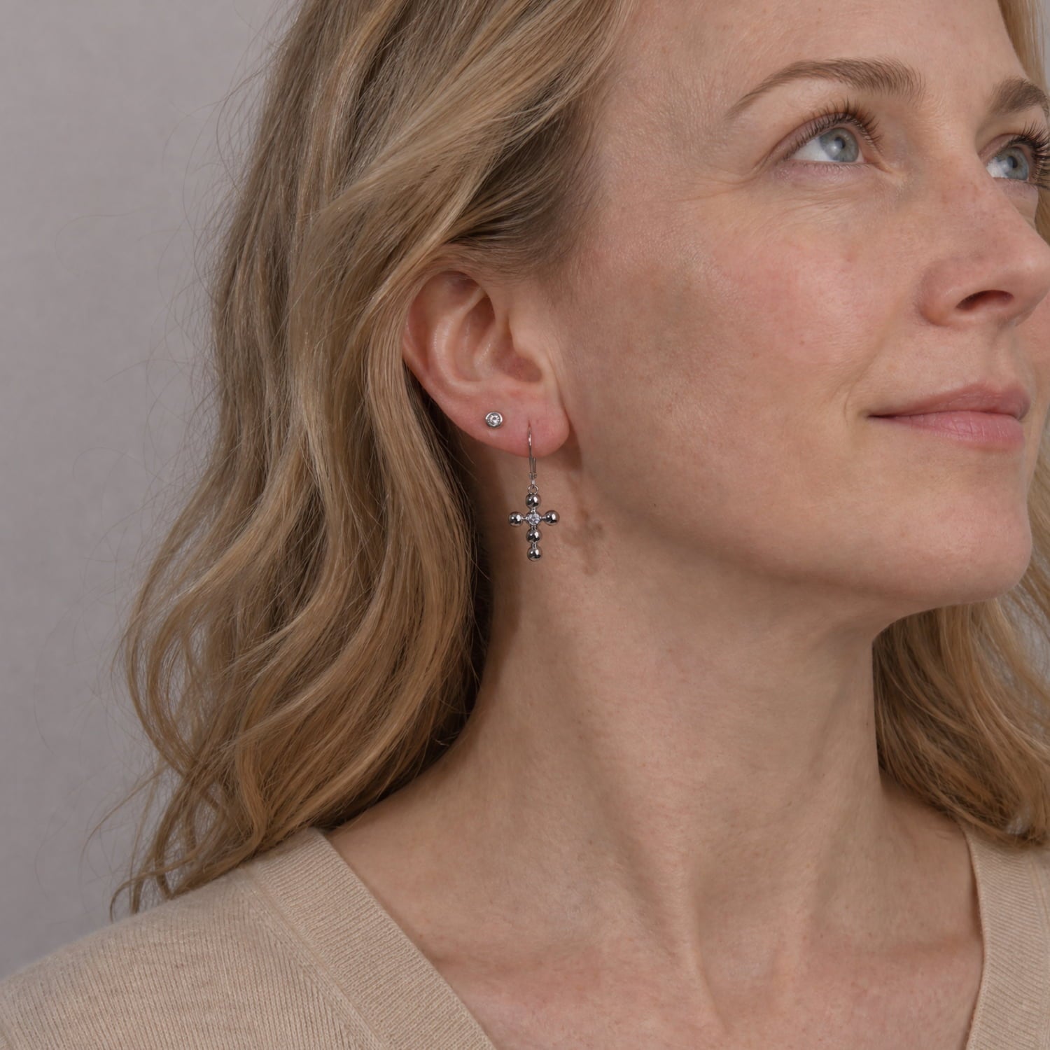 A close-up of a woman wearing a silver drop earring with a small cross and clear stones, paired with a matching stud earring, against a neutral beige background.