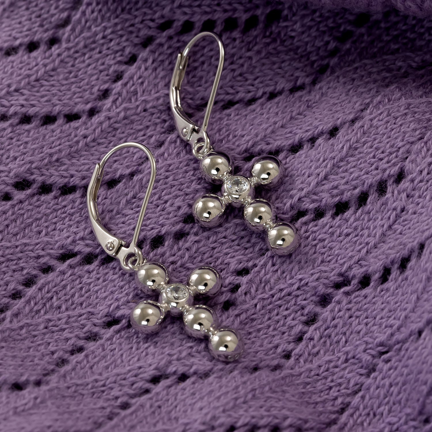 Pair of silver drop earrings featuring beaded cross pendants accented with central cubic zirconia stones, displayed on a textured purple fabric background.