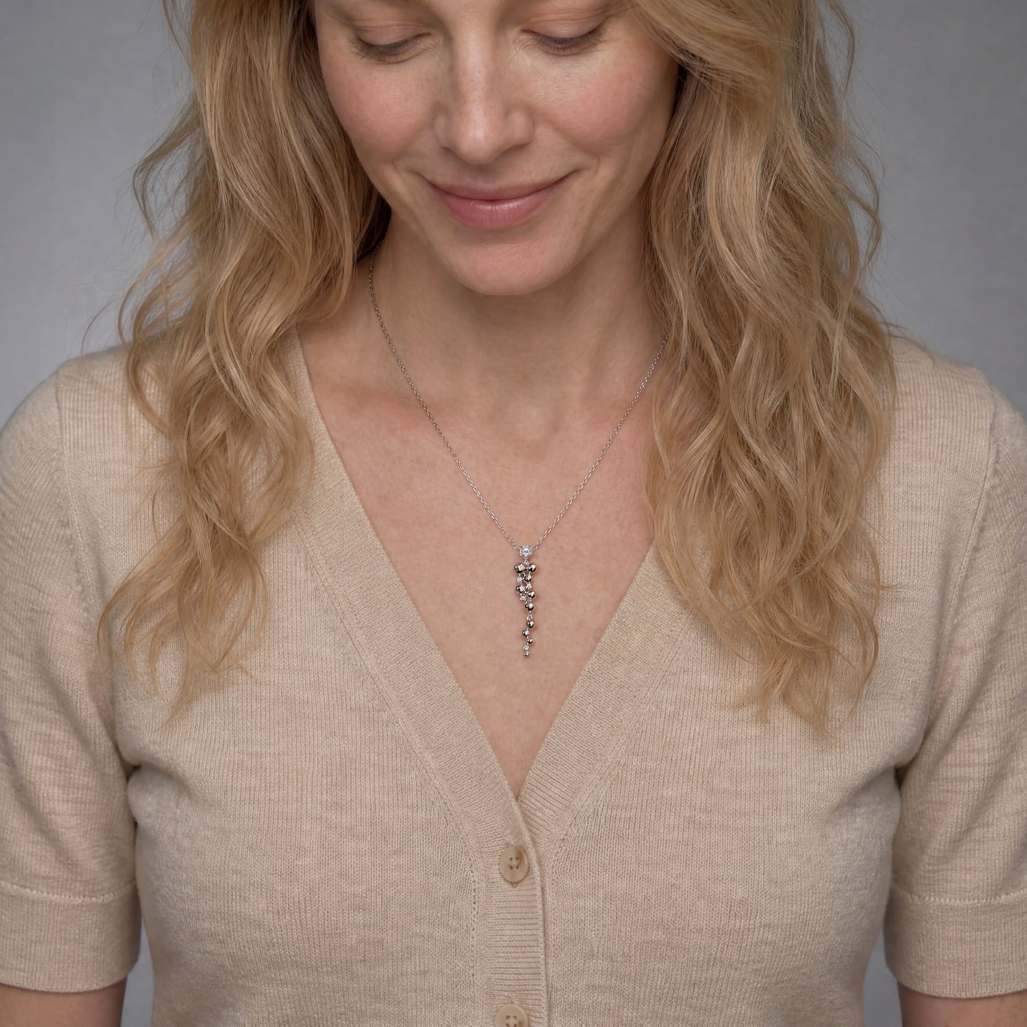 A woman with blonde wavy hair wearing a beige V-neck cardigan and a delicate silver waterfall necklace shown against a soft gray background.