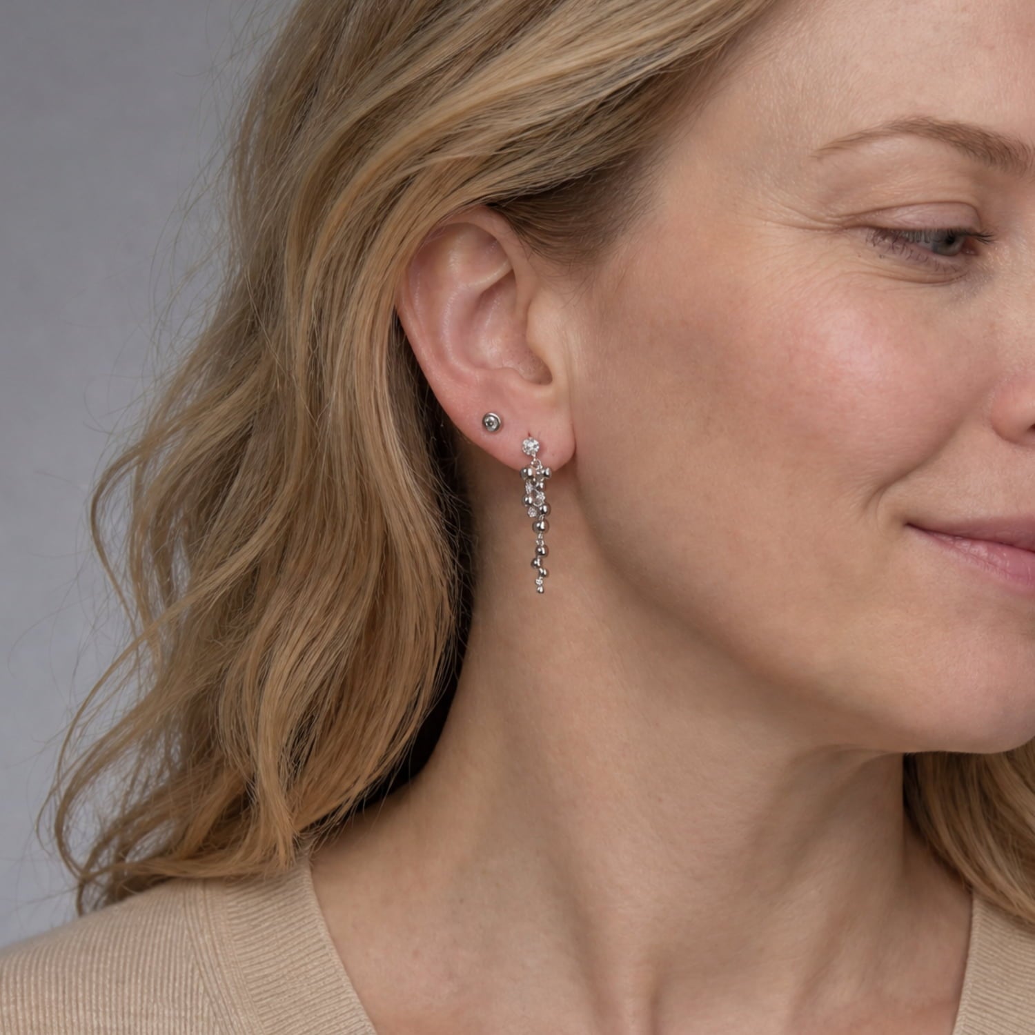A close-up of a woman's ear wearing a delicate silver dangle stud earring with small clear stones, alongside a matching small silver stud.