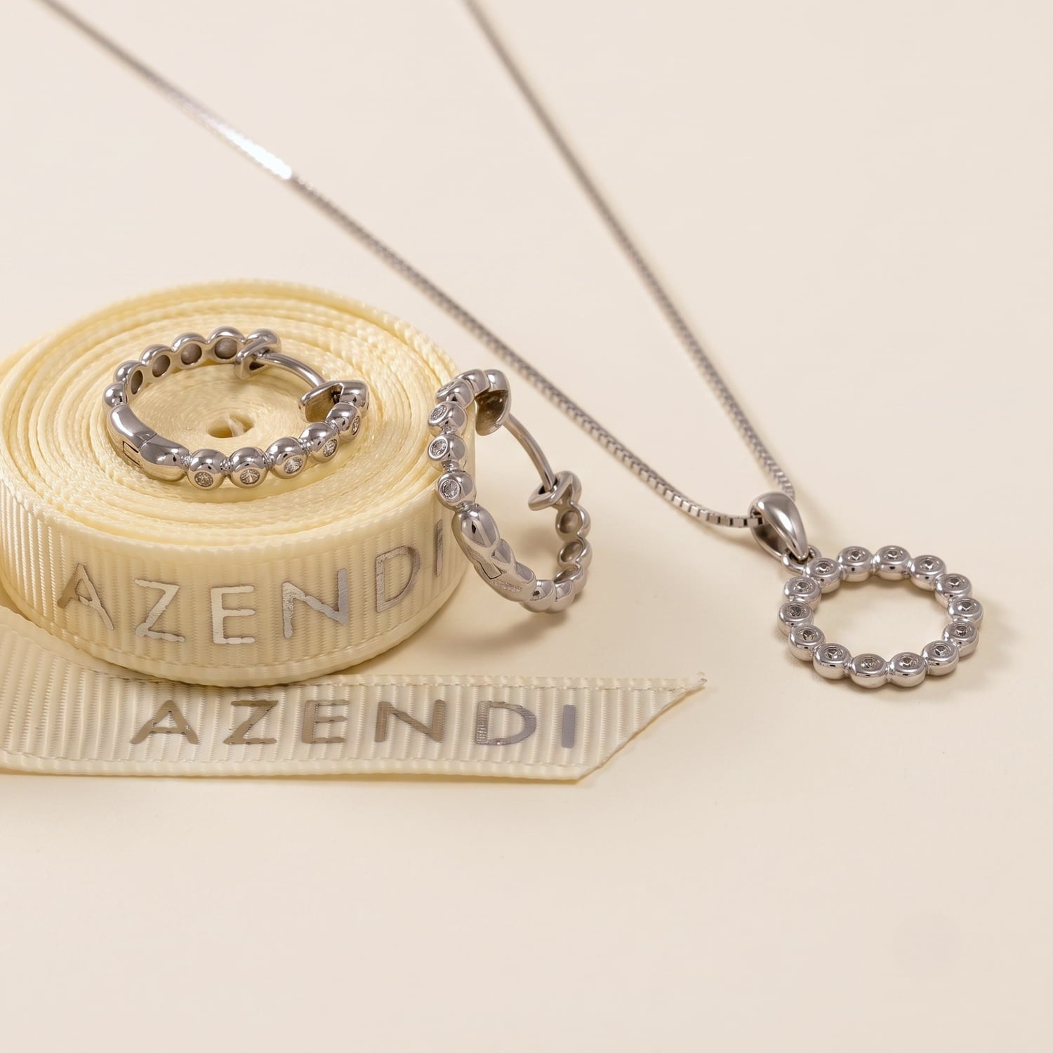 Sterling silver hoop earrings and matching pendant featuring a beaded design with cubic zirconia accents, displayed on a wooden surface alongside Azendi branded ribbon.