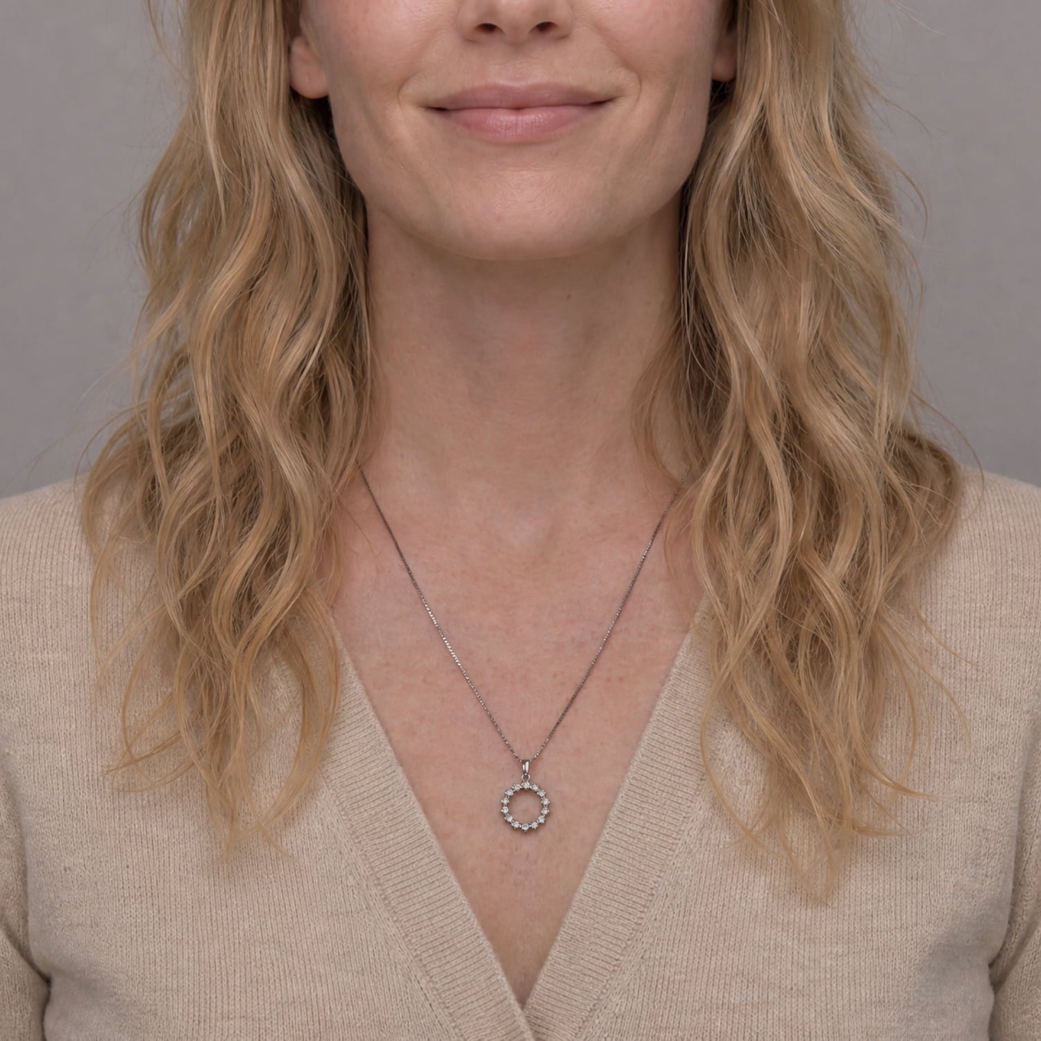 A close-up of a woman with blonde wavy hair wearing a delicate silver necklace with a small circular pendant, paired with a beige V-neck sweater.