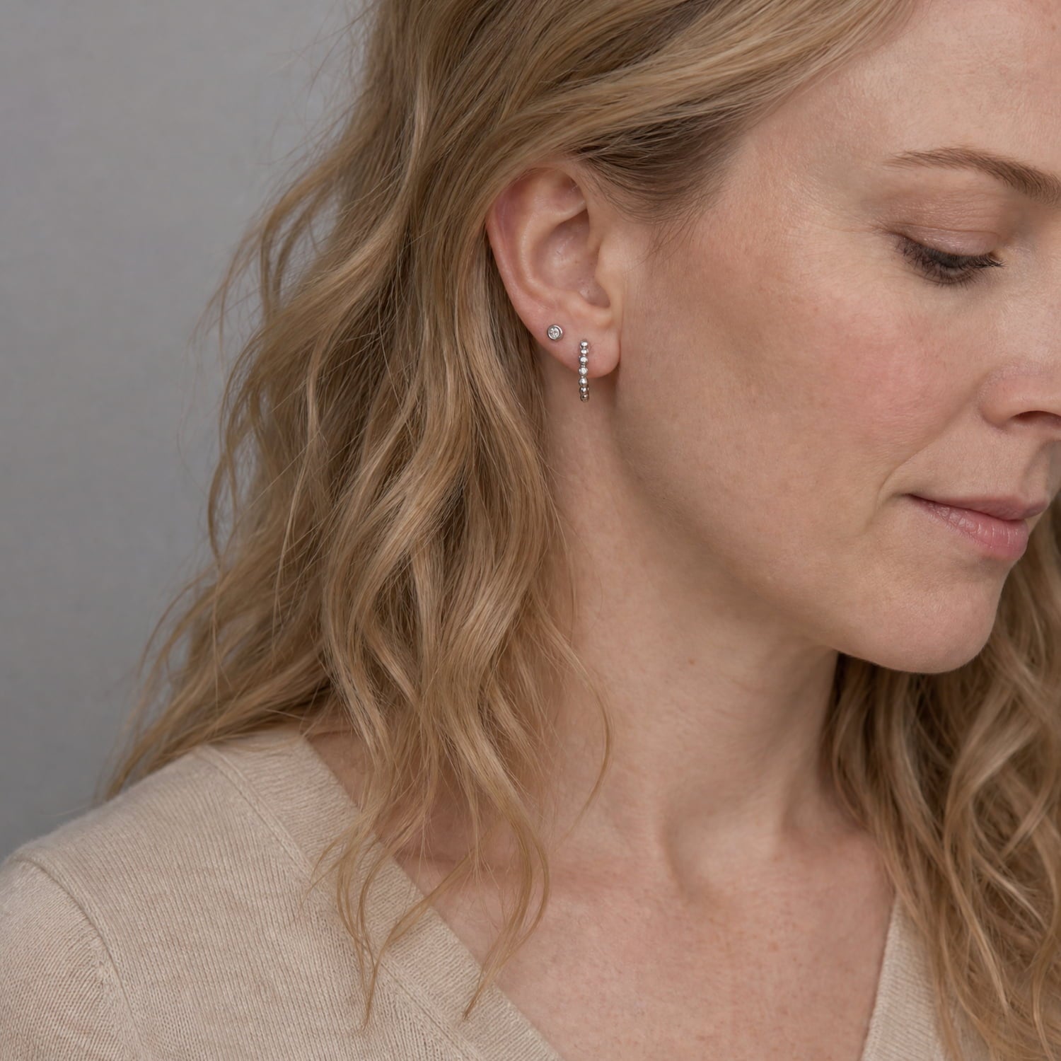 A close-up of a woman with blonde hair wearing a pair of small silver hoop earrings with tiny clear stones, shown against a neutral gray background.