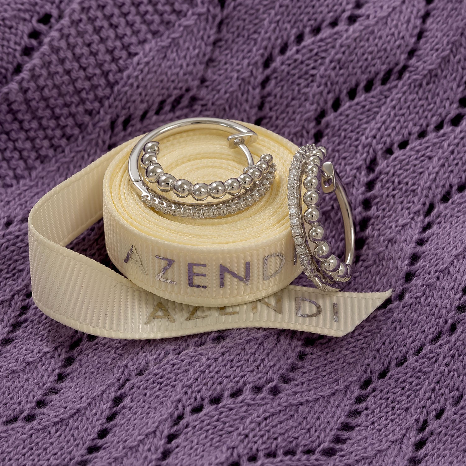 Pair of sterling silver double hoop earrings featuring a row of polished beads and cubic zirconia accents, displayed on an Azendi-branded cream ribbon against textured purple fabric.