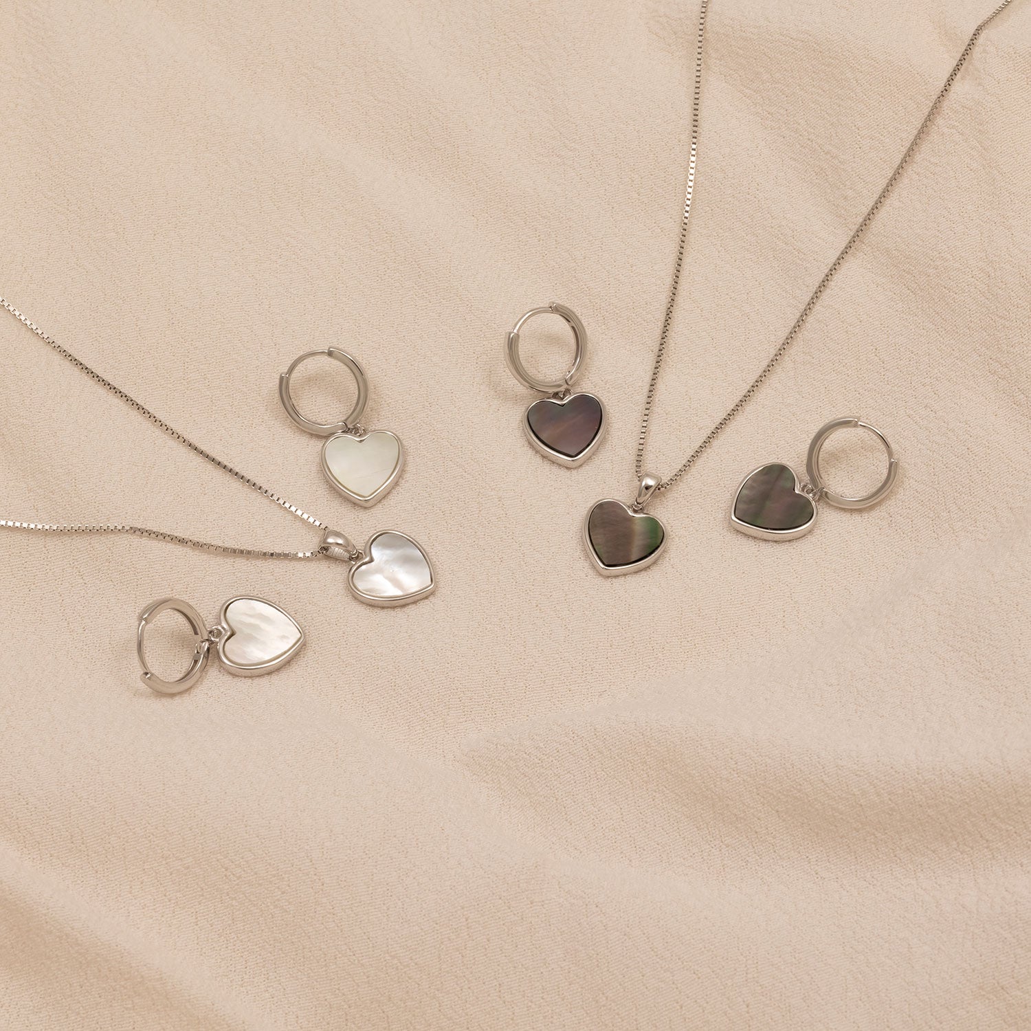 Collection of sterling silver heart charm hoop earrings and matching necklaces featuring grey and white mother of pearl inlays, displayed on a soft beige fabric background.