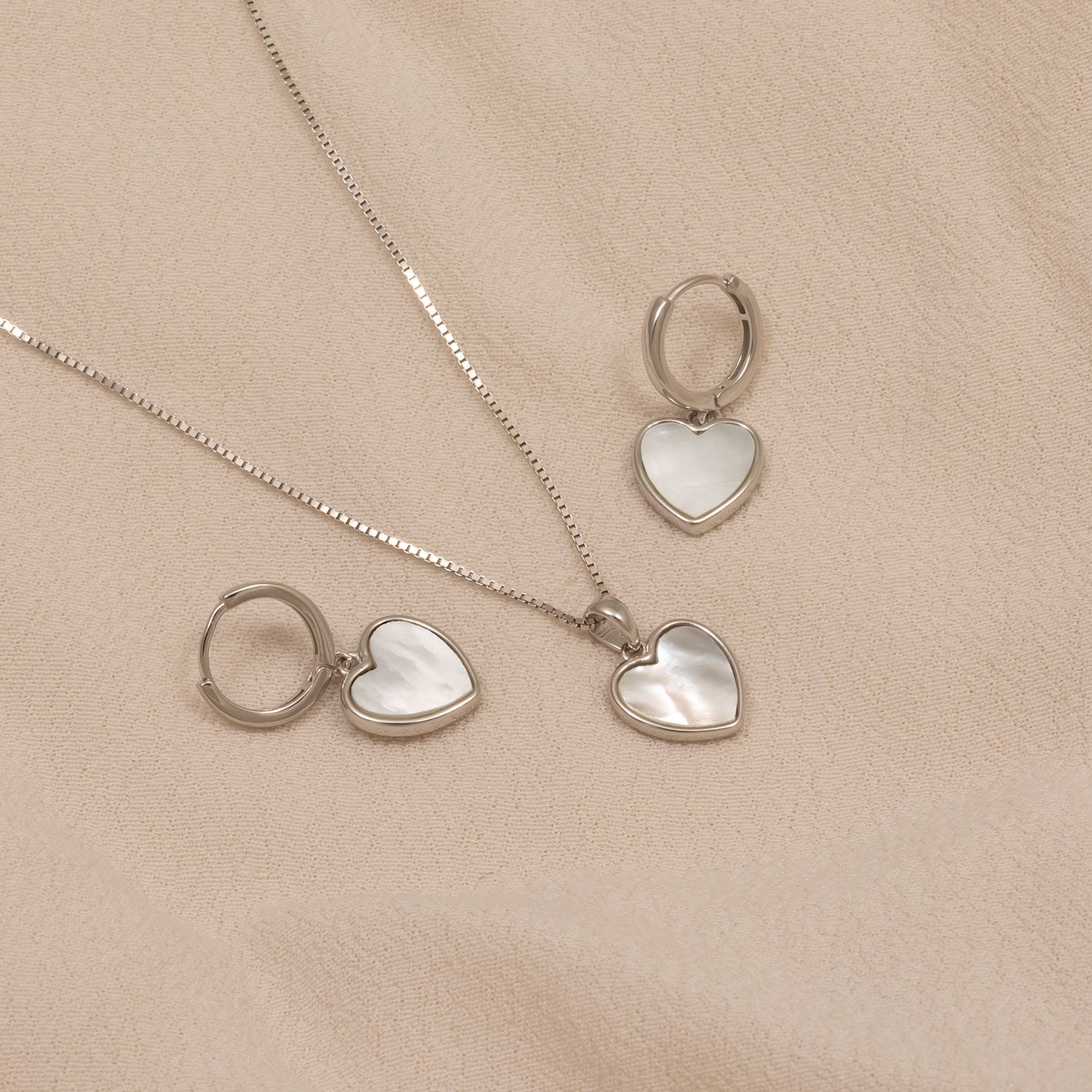 Pair of sterling silver hoop earrings and matching pendant necklace featuring white mother of pearl heart-shaped charms, styled on a soft beige fabric background.