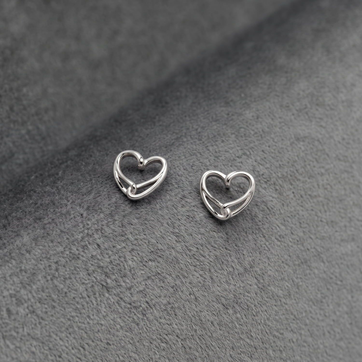A pair of small silver heart-shaped stud earrings on a textured dark gray fabric background.