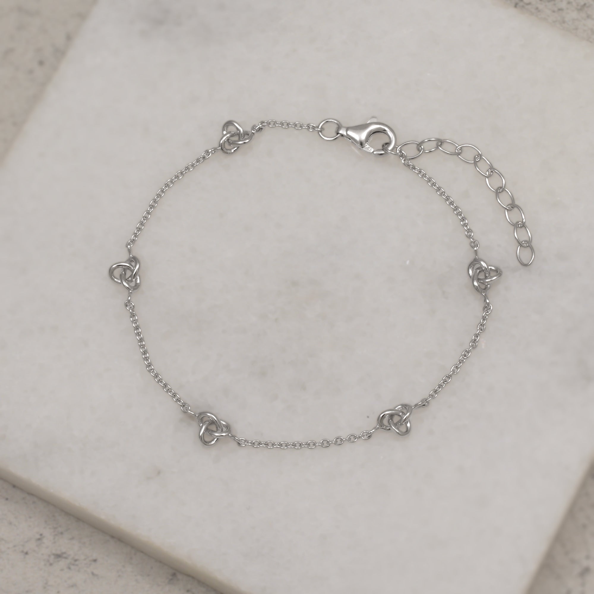 Sterling silver bracelet featuring delicate interconnected knot motifs along a fine chain with an adjustable extension for a subtle and refined contemporary jewellery look. Placed on a white quartz tile.