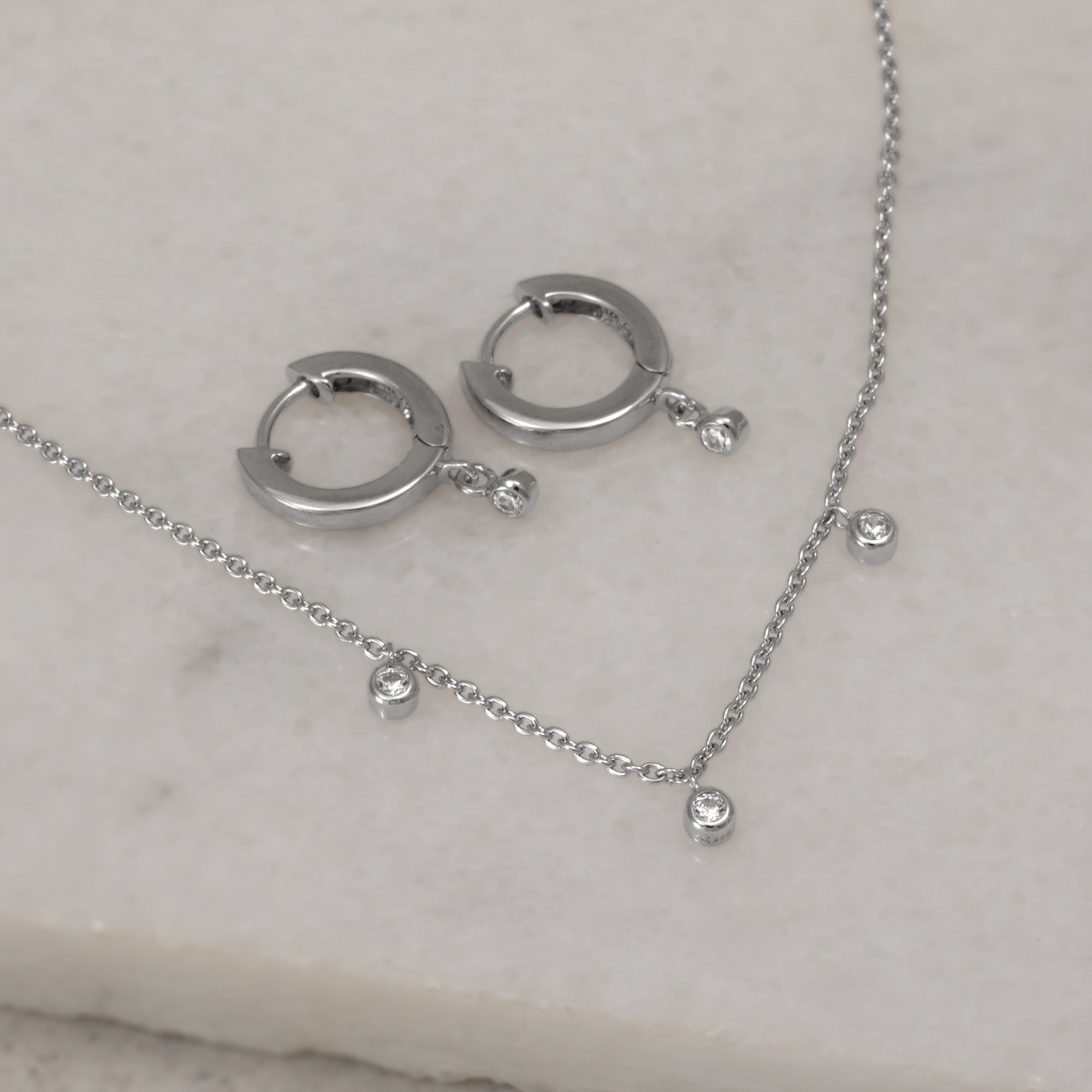 Delicate sterling silver necklace featuring three small gemstone accents suspended along a fine chain with an adjustable extension for versatile wear. Sterling silver hinged hoop earrings featuring a delicate hanging sparkling stone. Placed on a white quart tile.