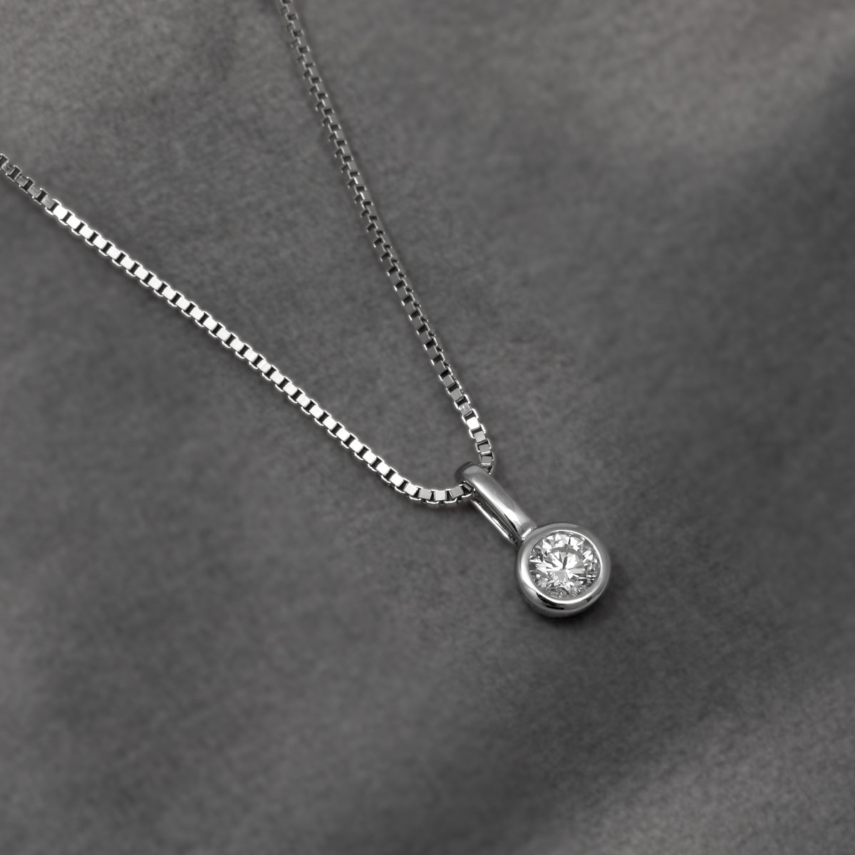 Silver necklace with a round bezel-set diamond pendant, shown on a dark gray textured background.