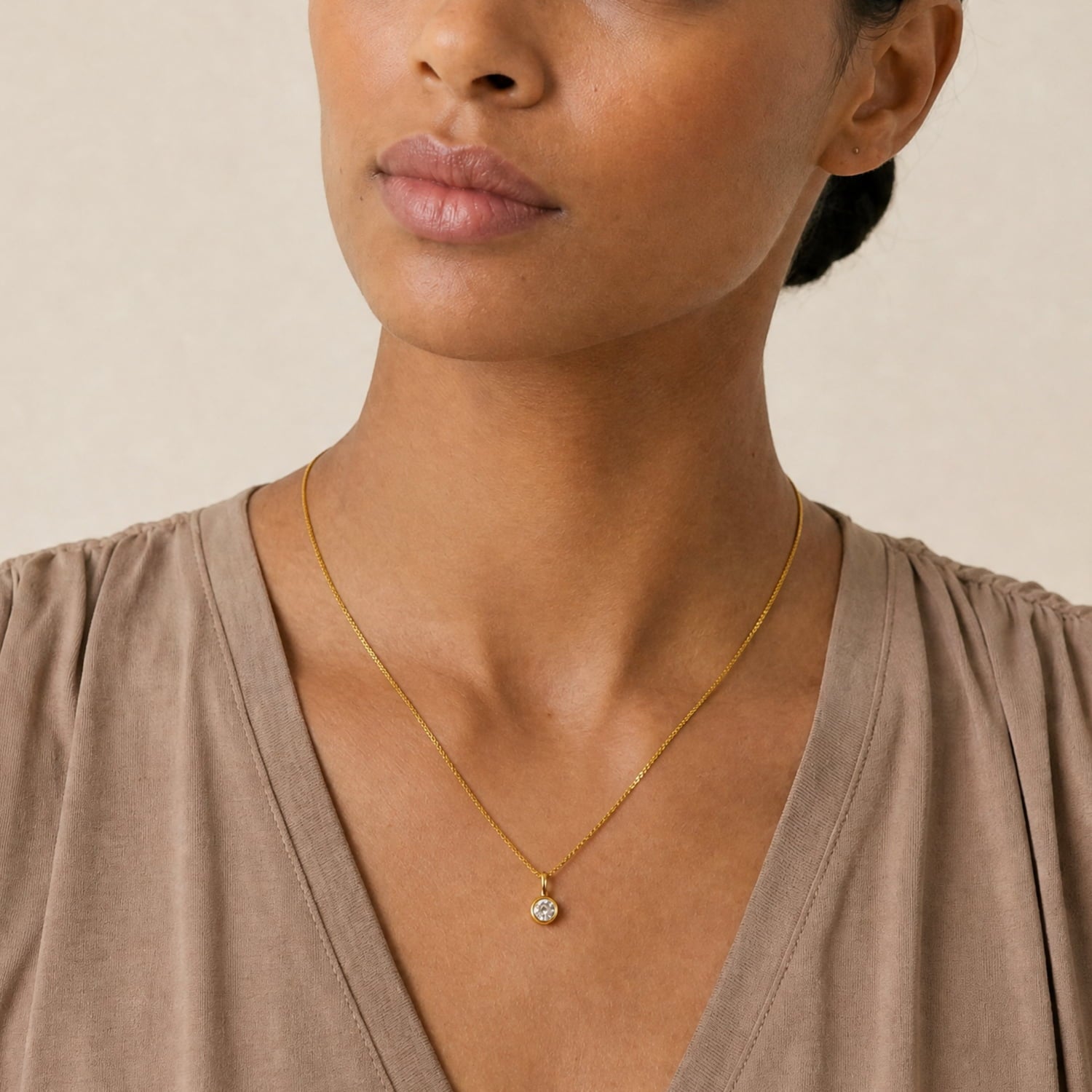 A close-up of a model wearing a delicate gold vermeilchain necklace with a round lab grown diamond pendant, paired with a taupe brown V-neck top against a neutral beige background.