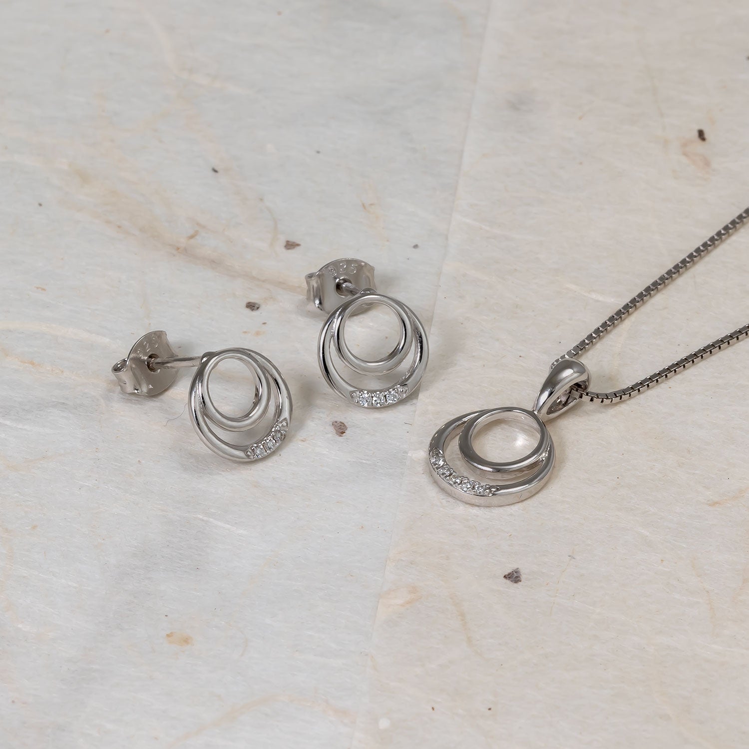 Sterling silver stud earrings featuring two interlocking circles accented with lab grown diamonds, paired with a matching pendant necklace on a light stone surface.