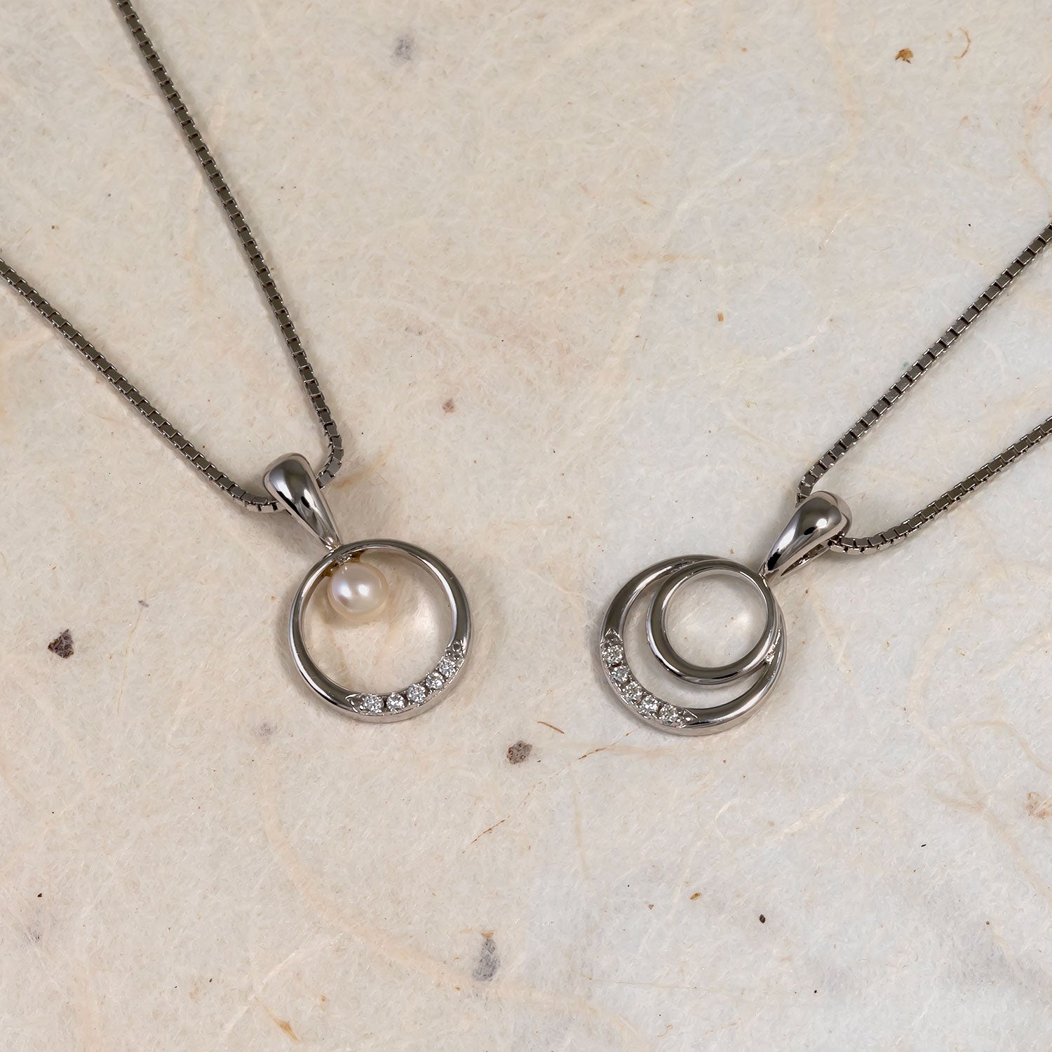Two sterling silver pendant necklaces featuring circular designs accented with small lab grown diamonds; one includes a freshwater pearl suspended inside the open circle.