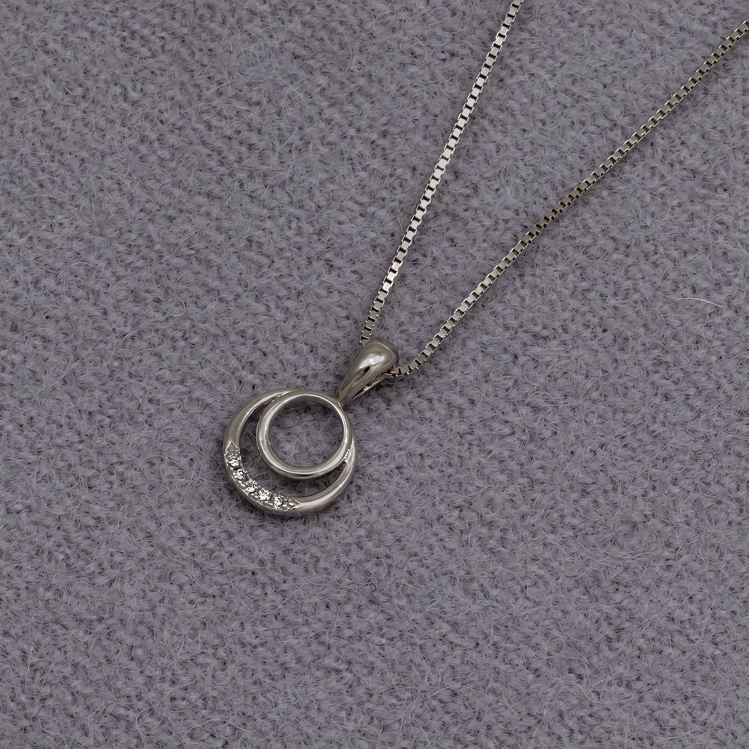 Sterling silver pendant featuring two interlocking circles, one accented with 0.02 carat lab grown diamonds, suspended from a fine box chain on a soft grey textured background.