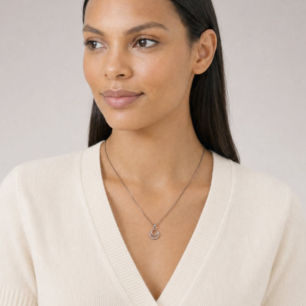 A woman wearing a delicate silver and lab diamond pendant, styled with a cream-colored V-neck sweater against a neutral light gray background.