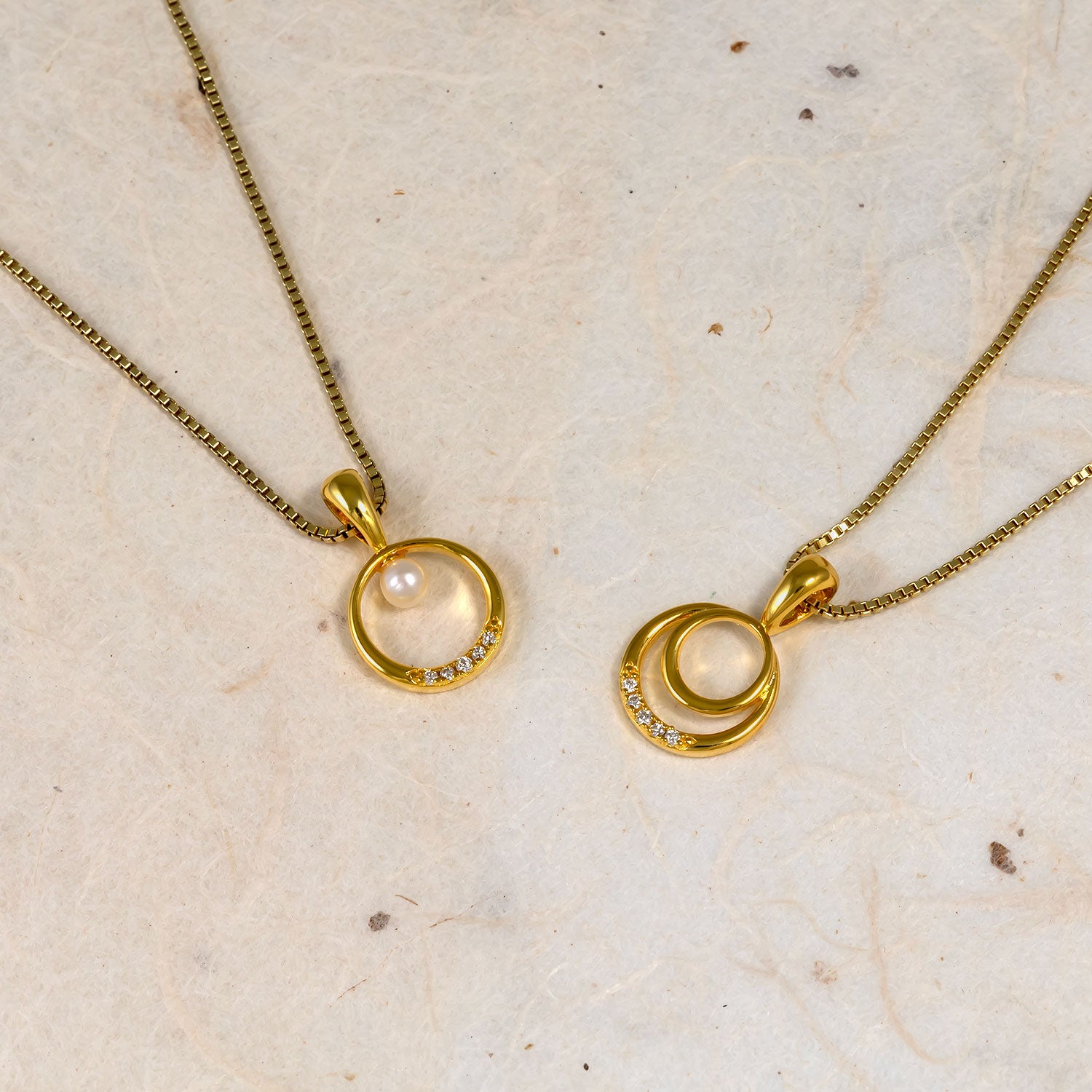 Two gold vermeil pendants featuring overlapping circular designs accented with small lab grown diamonds and a single freshwater pearl, displayed on a neutral stone surface.