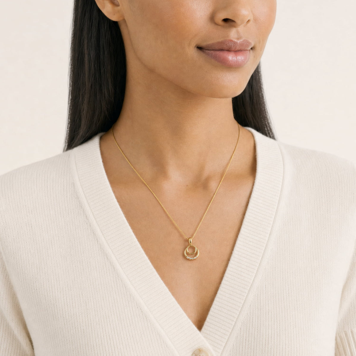 A close-up of a woman wearing a delicate gold vermeil chain necklace with a small interlocking rings pendant, styled with a cream ribbed V-neck sweater.