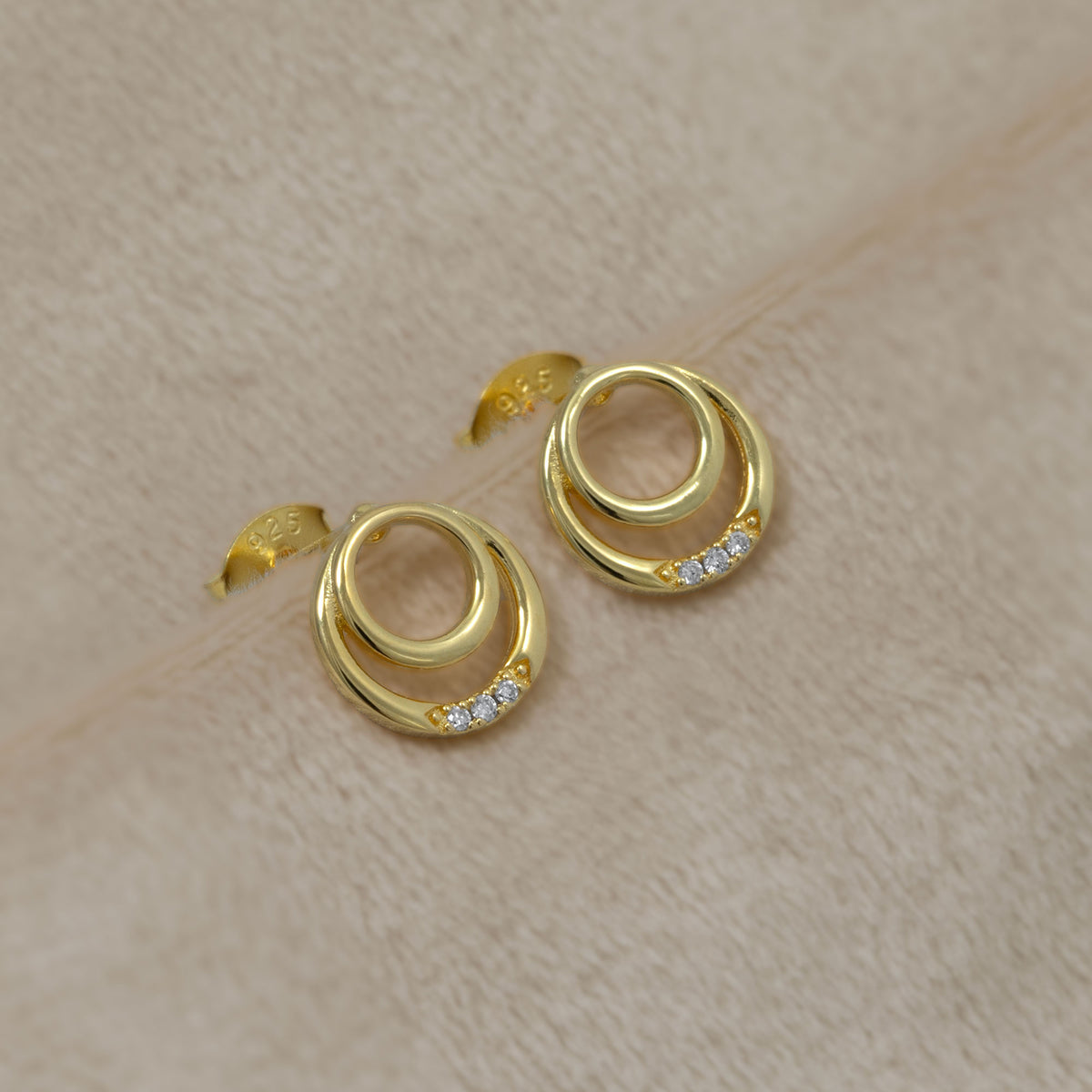 Delicate gold vermeil stud earrings featuring overlapping double circles accented with a row of sparkling lab grown diamonds, set against a soft cream textured background.