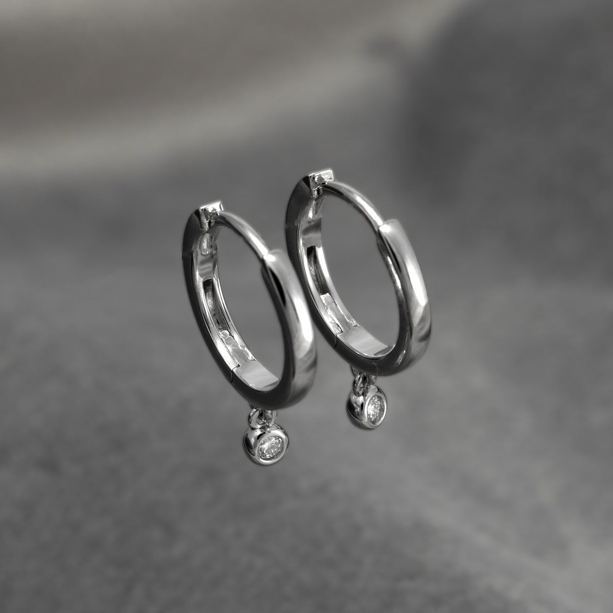Hinged sterling silver hoops featuring small round charms each set with a 0.05 carat lab grown diamond, resting on a soft grey textured fabric background.