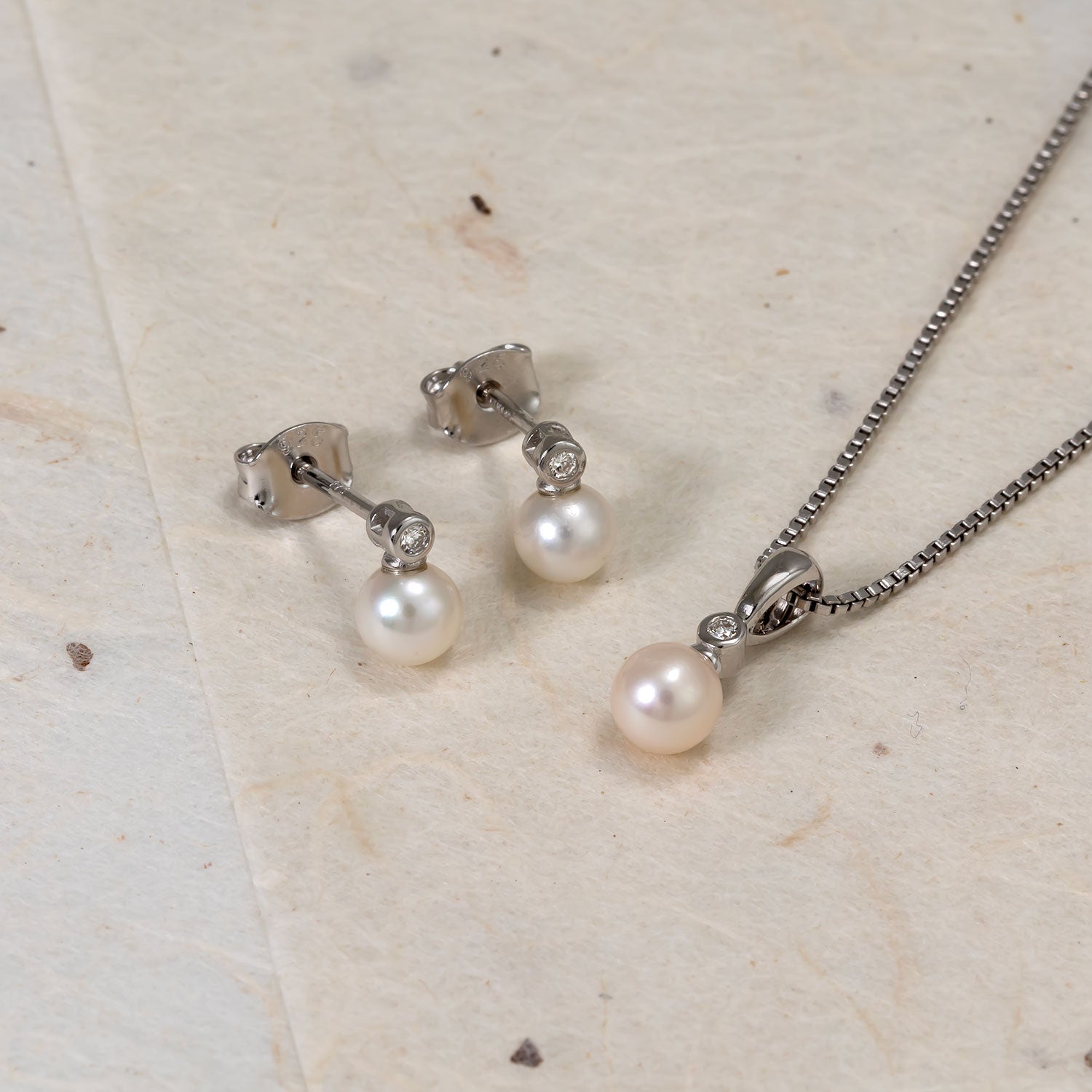 Pair of sterling silver stud earrings featuring freshwater pearls and small lab grown diamonds, displayed alongside a matching pearl pendant on a fine silver chain.