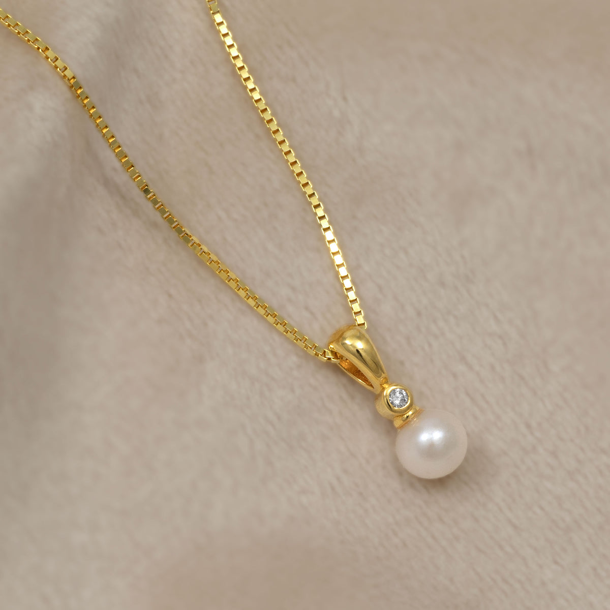Elegant gold vermeil pendant featuring a round freshwater pearl accented by a small sparkling lab grown diamond, suspended from a fine gold chain on a soft cream fabric.