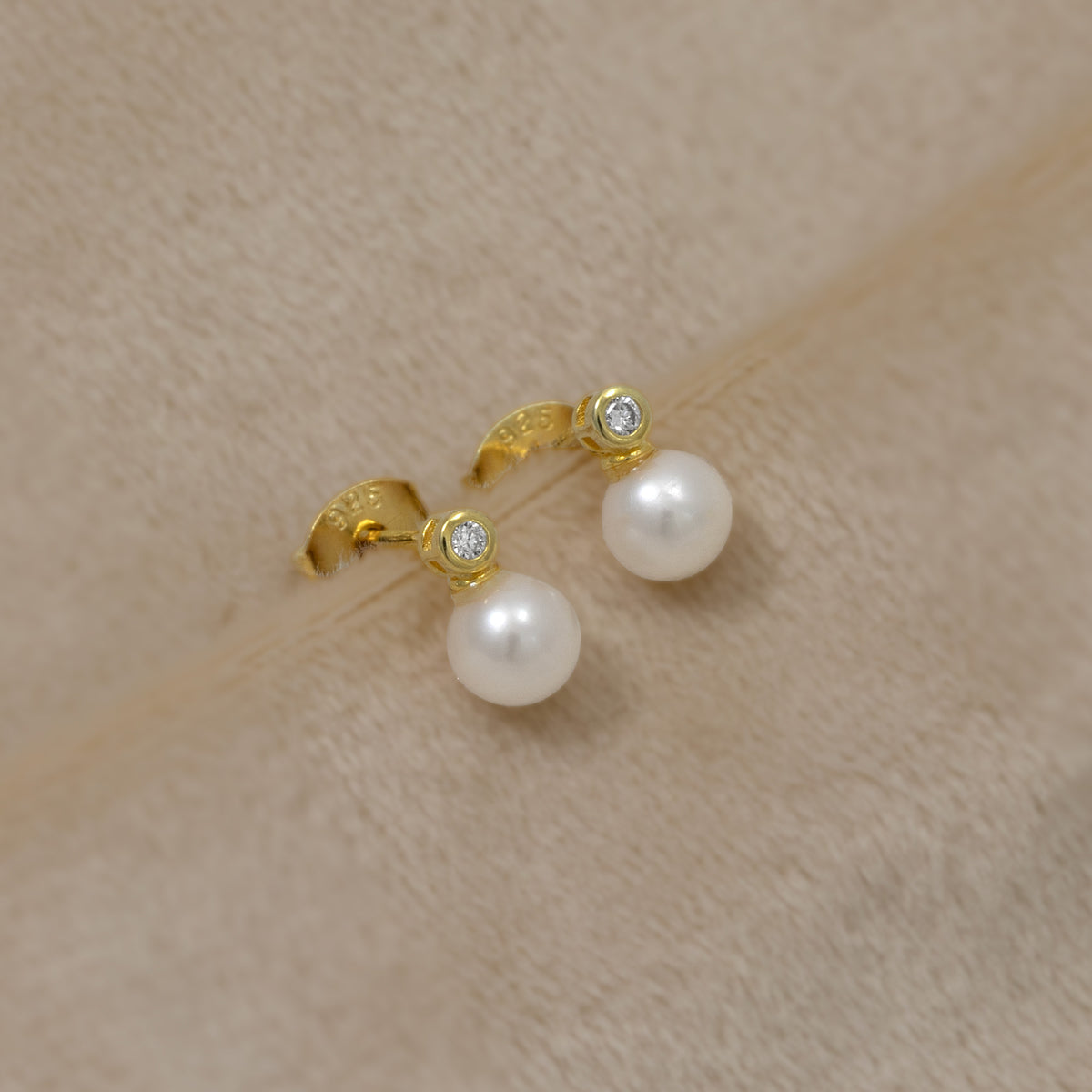 Pair of gold vermeil stud earrings featuring lustrous freshwater pearls accented with small lab grown diamonds, displayed on a soft cream textured fabric background.