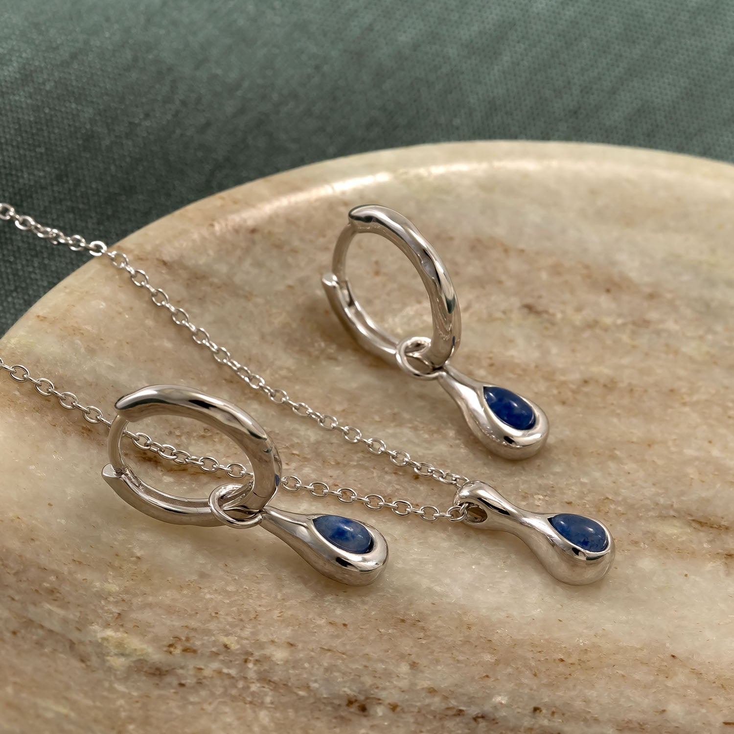 Sterling silver hoop earrings and matching necklace featuring teardrop-shaped blue aventurine stones, displayed on a smooth, neutral stone surface.