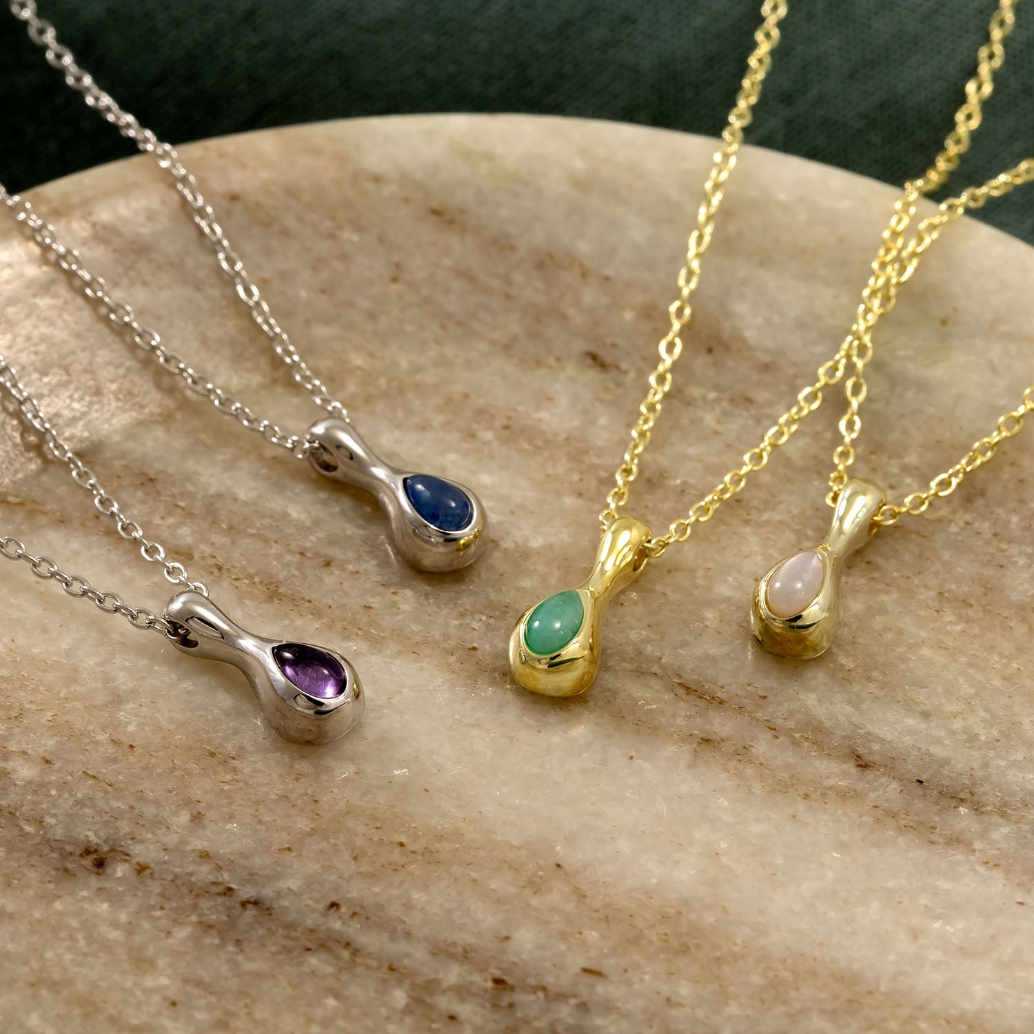 Collection of delicate necklaces featuring gold vermeil and sterling silver droplet pendants, each set with a different colored gemstone on fine chains displayed on a stone surface.