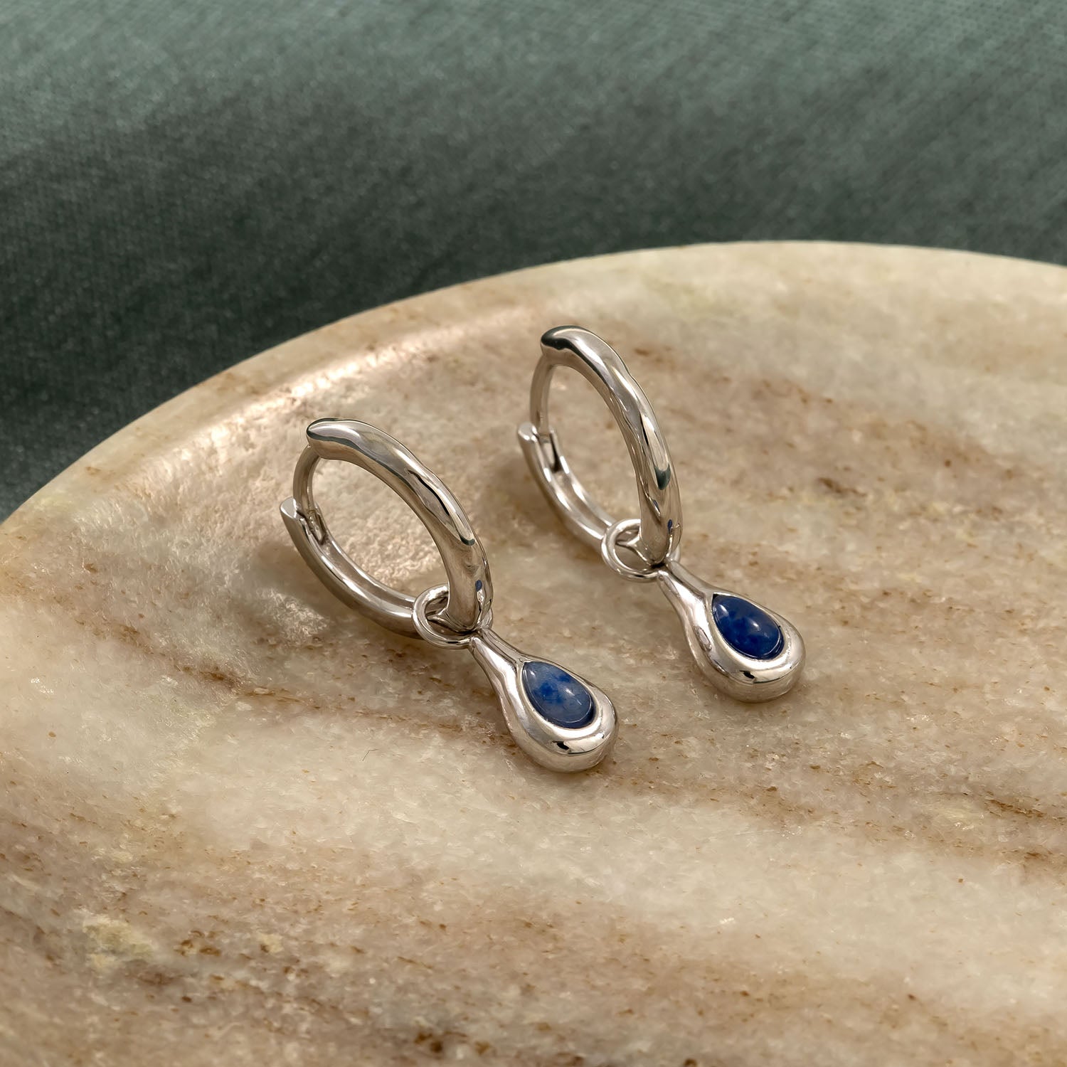 Pair of sterling silver hinge hoop earrings each featuring a teardrop-shaped blue aventurine charm, displayed on a smooth beige stone background.