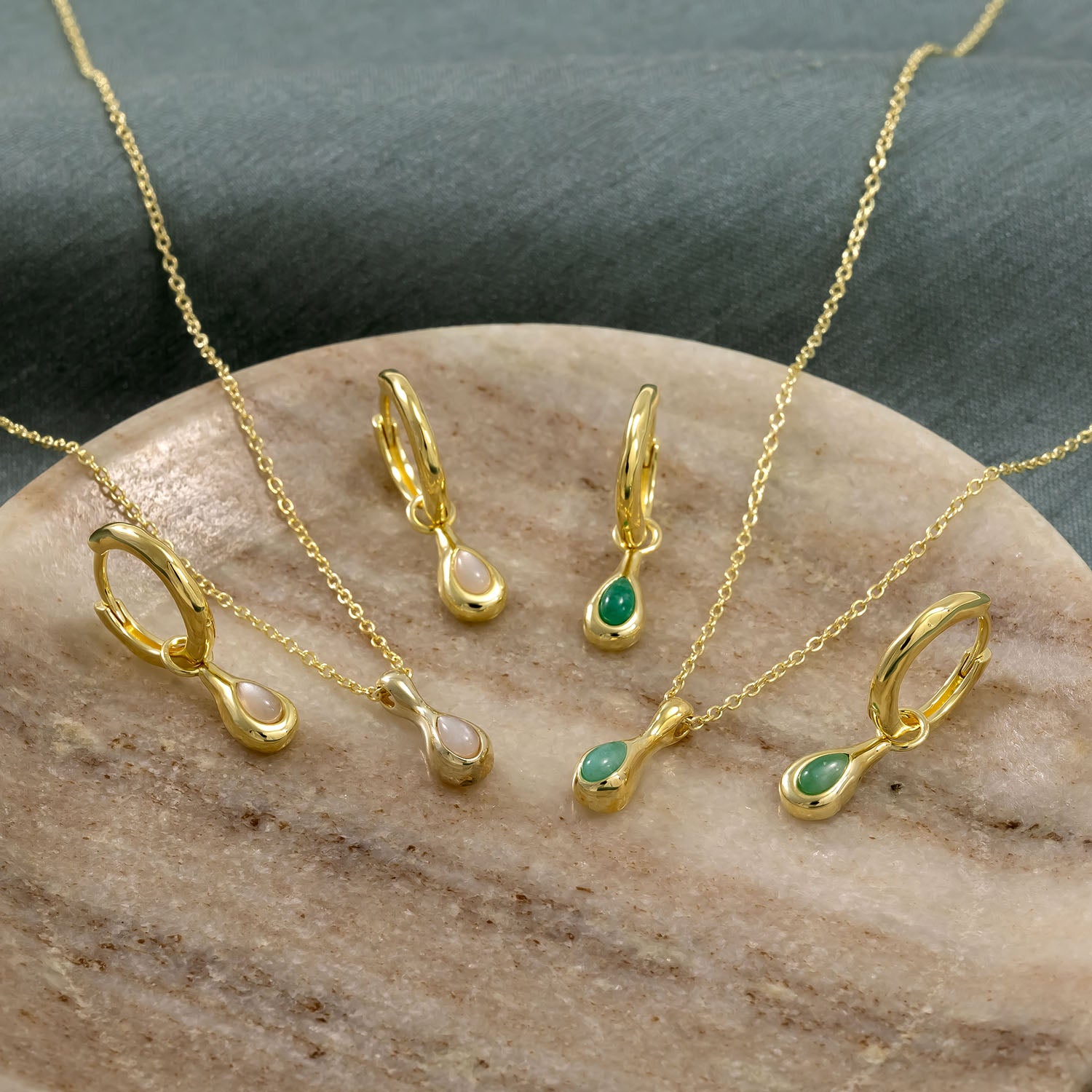 Two pairs of delicate gold vermeil hinge hoop earrings and matching pendant necklaces featuring smooth moonstone and green gemstone droplet charms, displayed on a neutral stone surface.