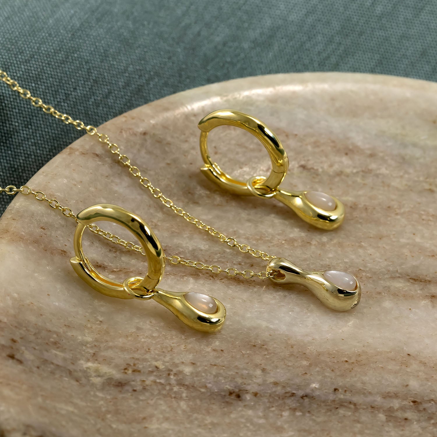 Pair of gold vermeil hinge hoop earrings each featuring a teardrop-shaped moonstone charm, displayed alongside a matching delicate chain necklace on a smooth stone surface.