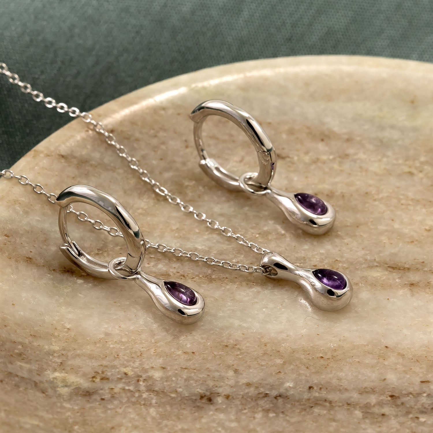 Pair of sterling silver hinge hoop earrings and matching pendant necklace, each featuring a smooth amethyst droplet charm resting on a natural stone surface.