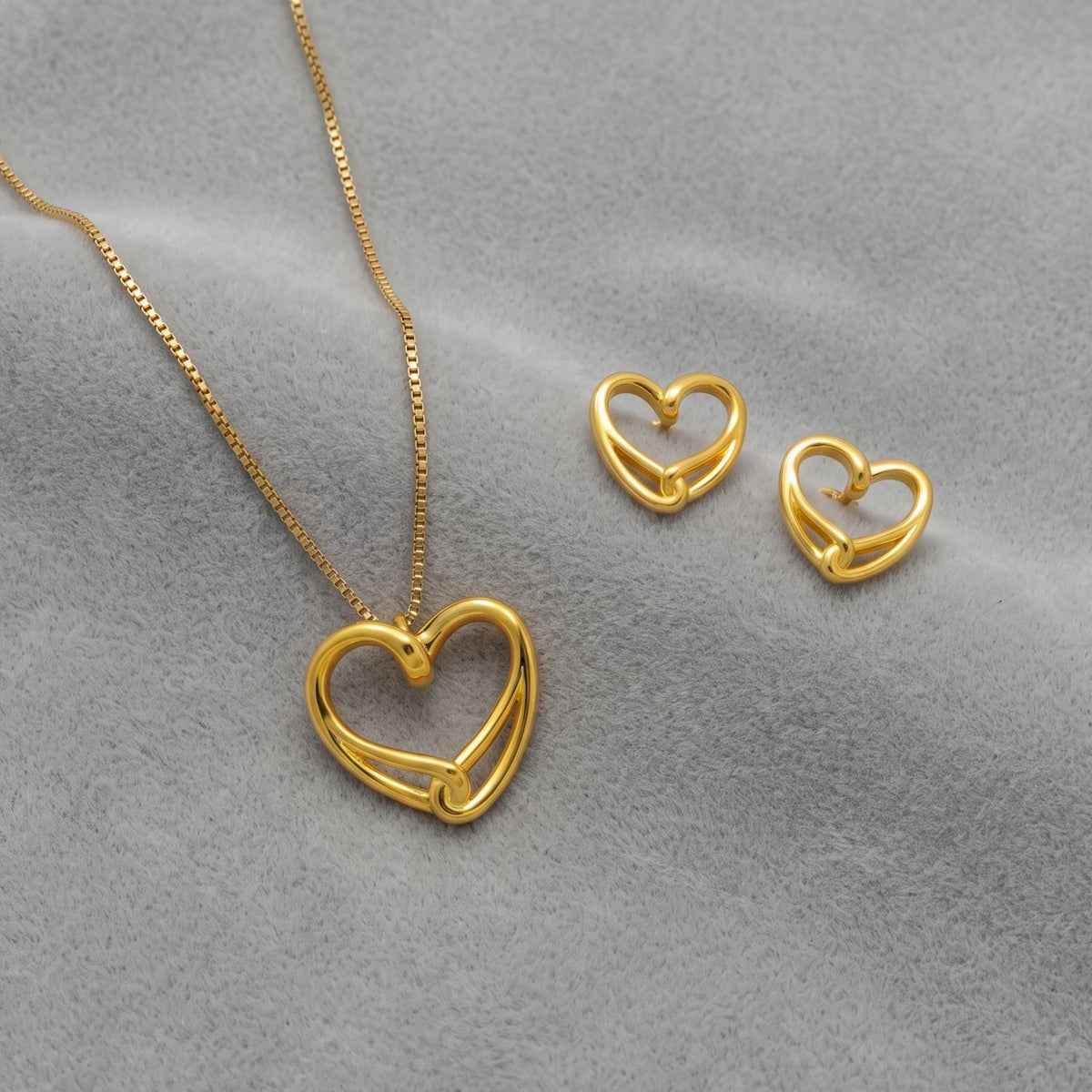 Gold vermeil heart-shaped pendant necklace and matching heart stud earrings displayed on a soft gray textured background.