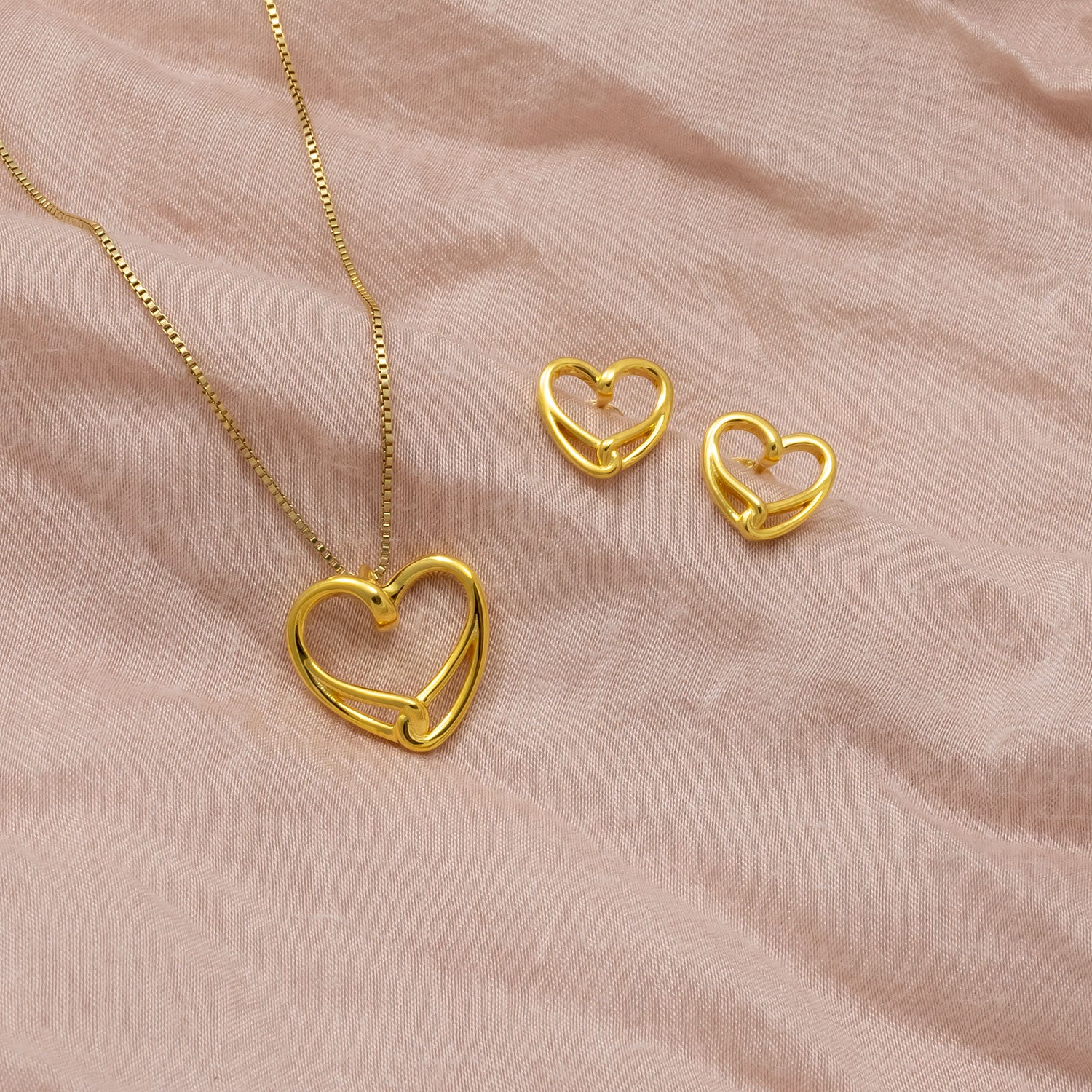 Set of gold vermeil heart-shaped stud earrings paired with a matching pendant necklace, featuring a modern intertwined design on a soft neutral fabric background.