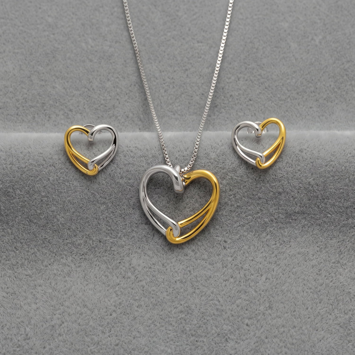 A pendant and stud earring set featuring a double-heart design in polished silver and yellow gold vermeil, displayed on a textured gray background.