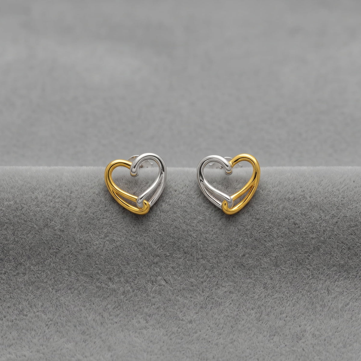 A pair of small heart-shaped stud earrings made of two-tone silver and gold vermeil, displayed on a textured gray surface.