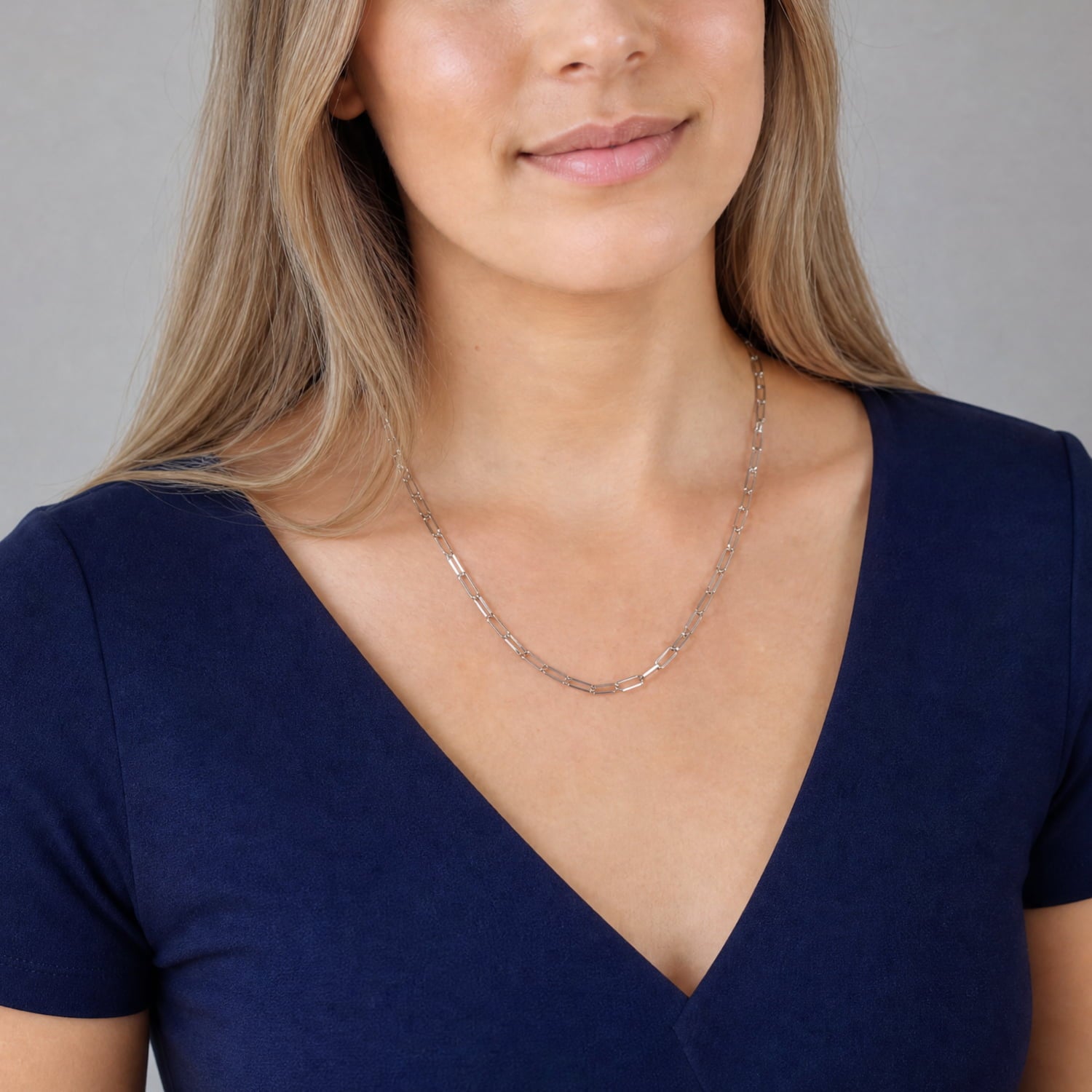 A close-up of a woman wearing a delicate silver chain necklace and a navy blue V-neck top, set against a plain grey background.