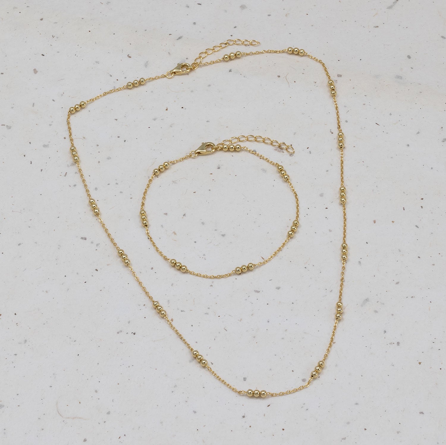 Delicate gold vermeil chain necklace and matching bracelet featuring groups of three small beads spaced evenly along fine chains, displayed on a neutral speckled surface.
