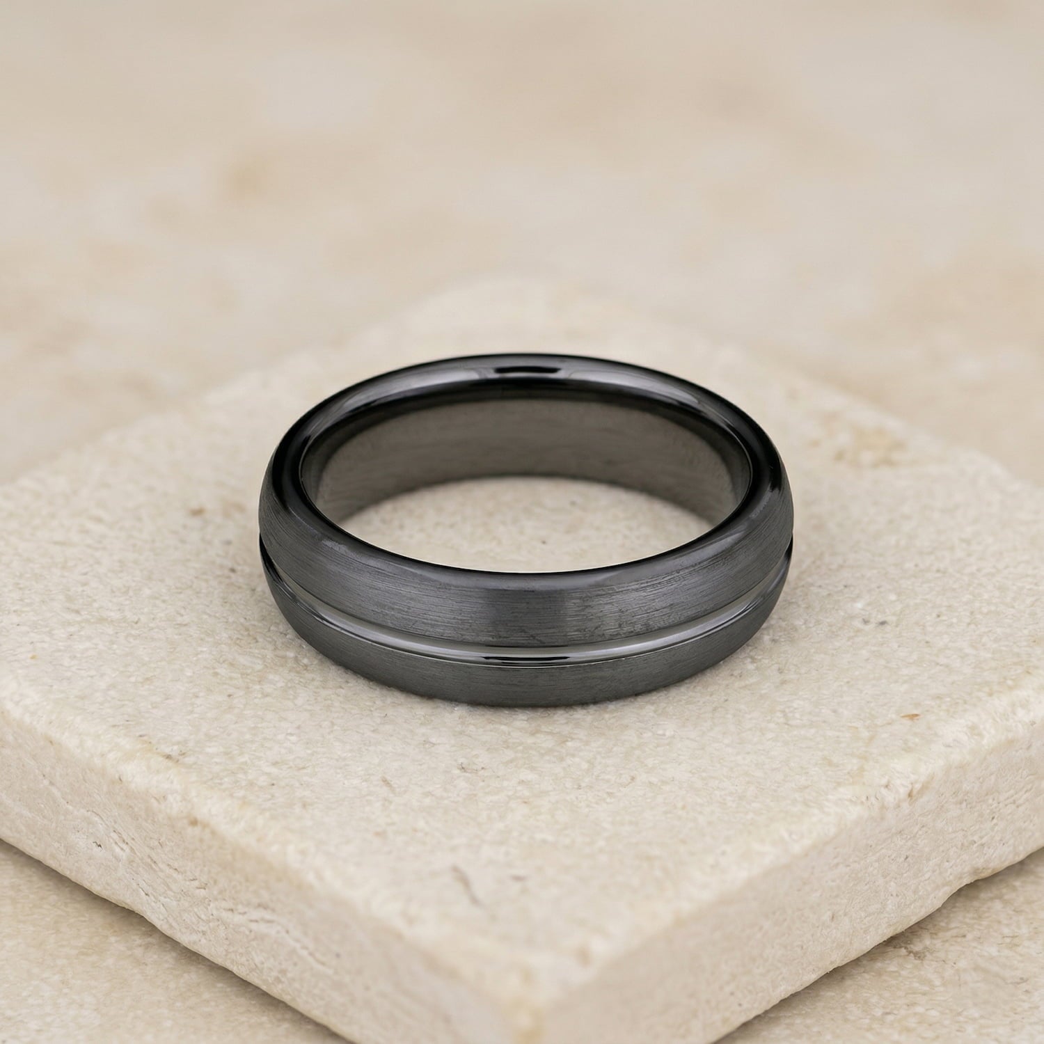 Polished tungsten ring featuring a smooth, central groove set against a matte finish, displayed on a textured neutral stone surface.